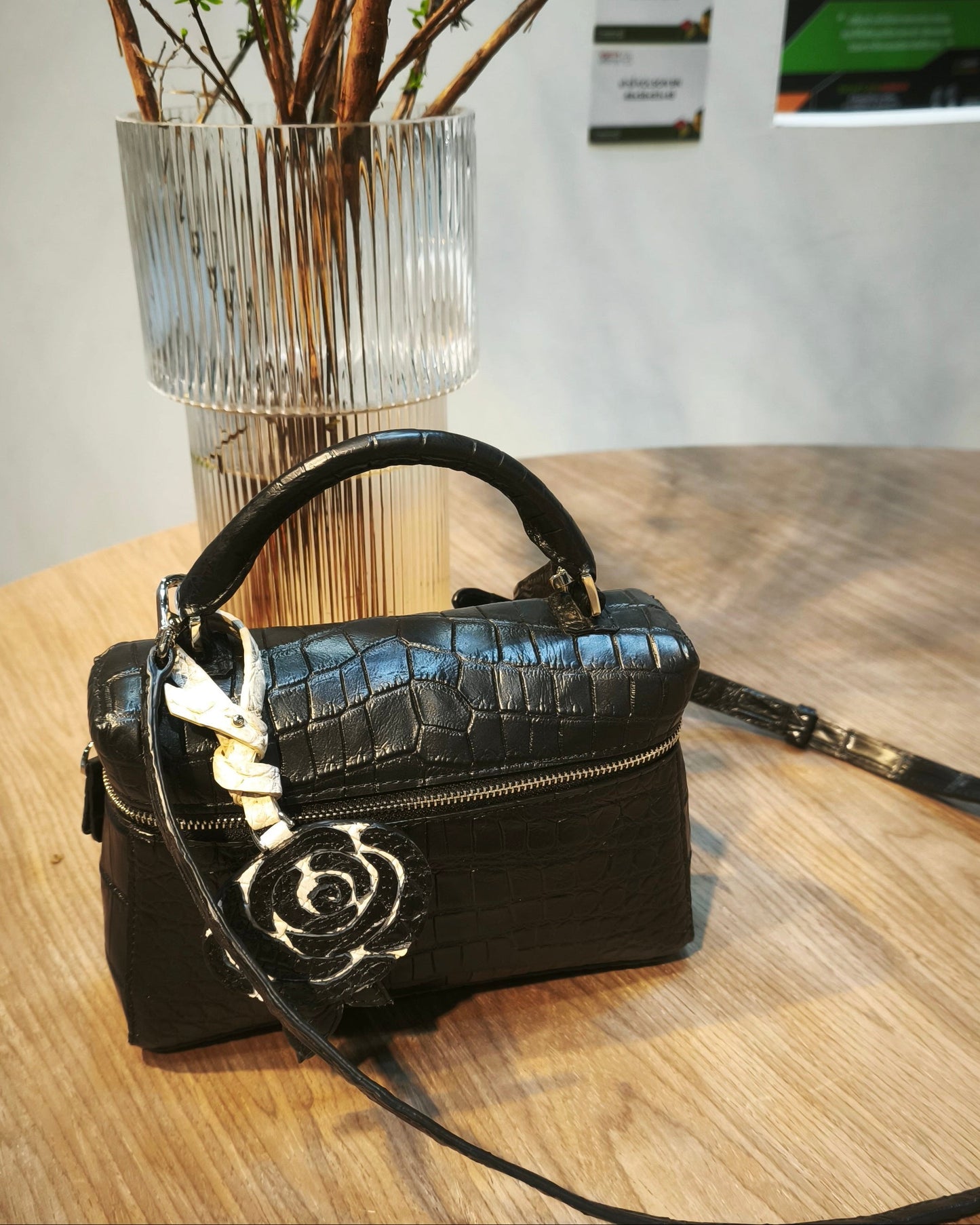 Black handbag with a floral clasp on a wooden table, with a vase of branches in the background.