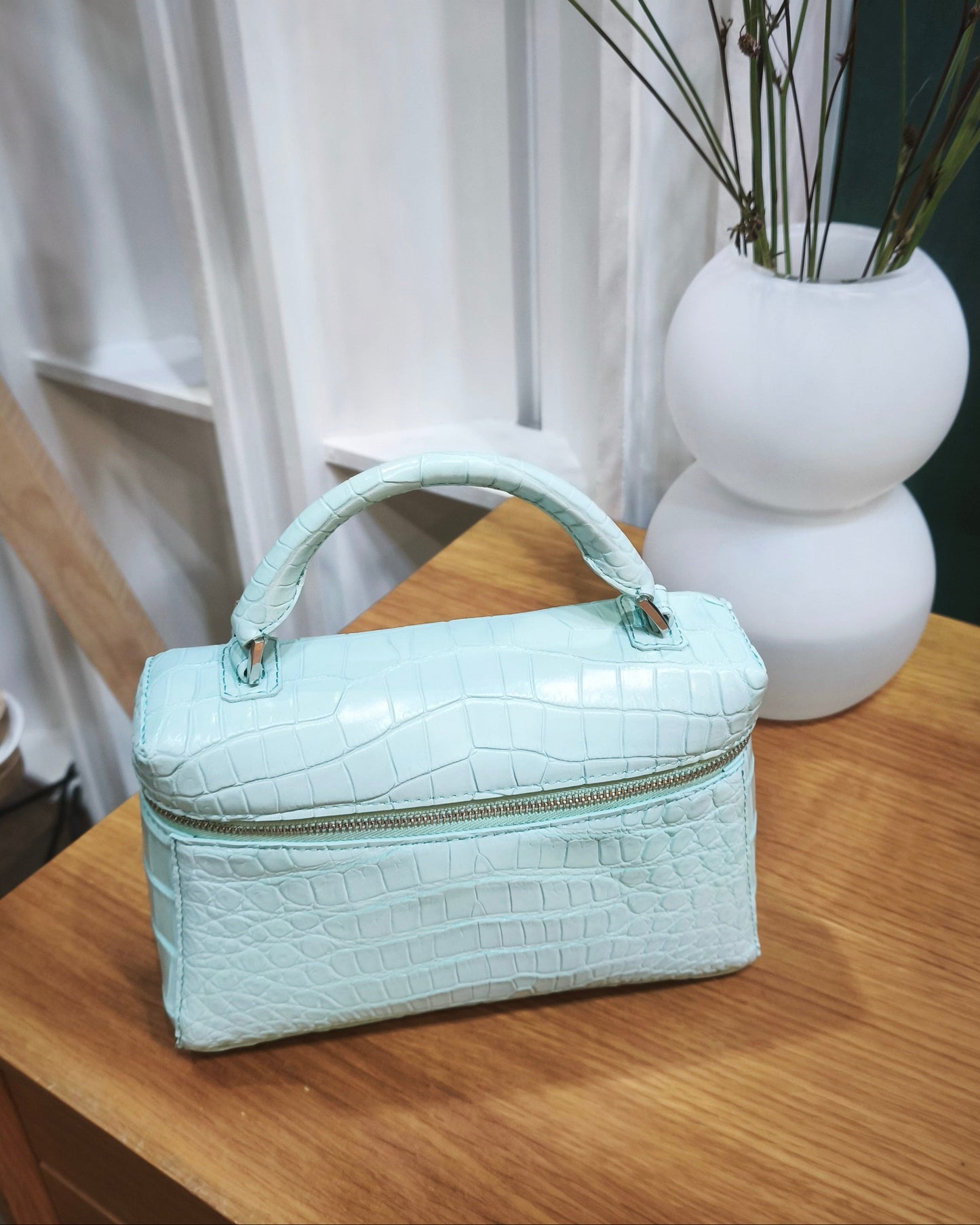 Light blue handbag on a wooden surface with a white vase and branches in the background.