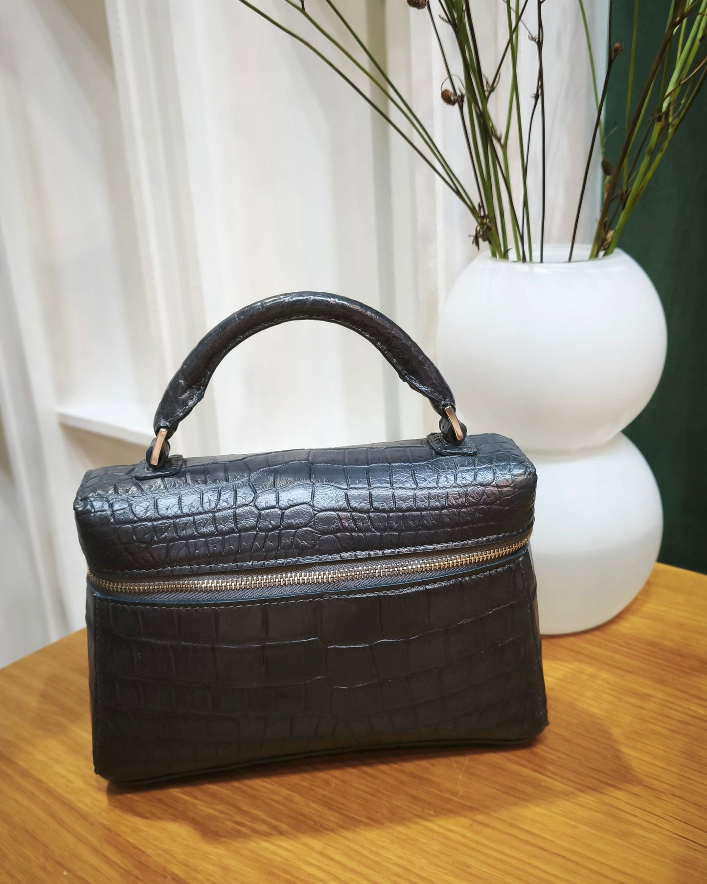Black handbag on a wooden surface with a white vase and branches in the background
