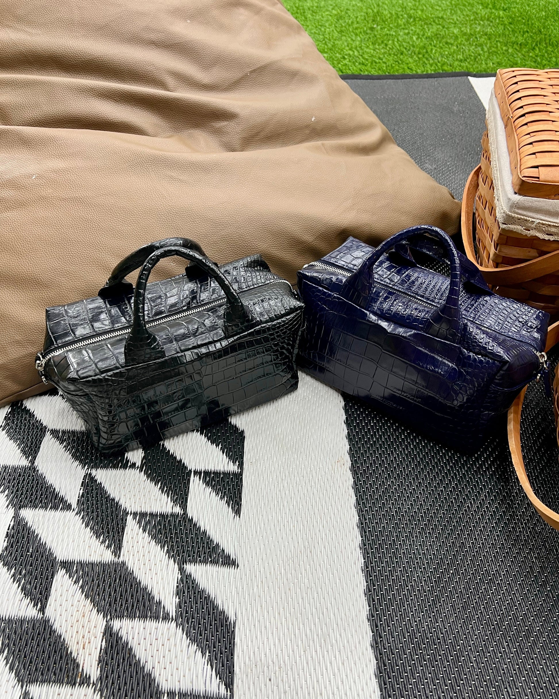 Two small structured handbags with a crocodile-embossed texture, one in glossy black and one in deep navy blue, displayed on a black and white geometric patterned rug next to a tan fabric bag and a woven basket.