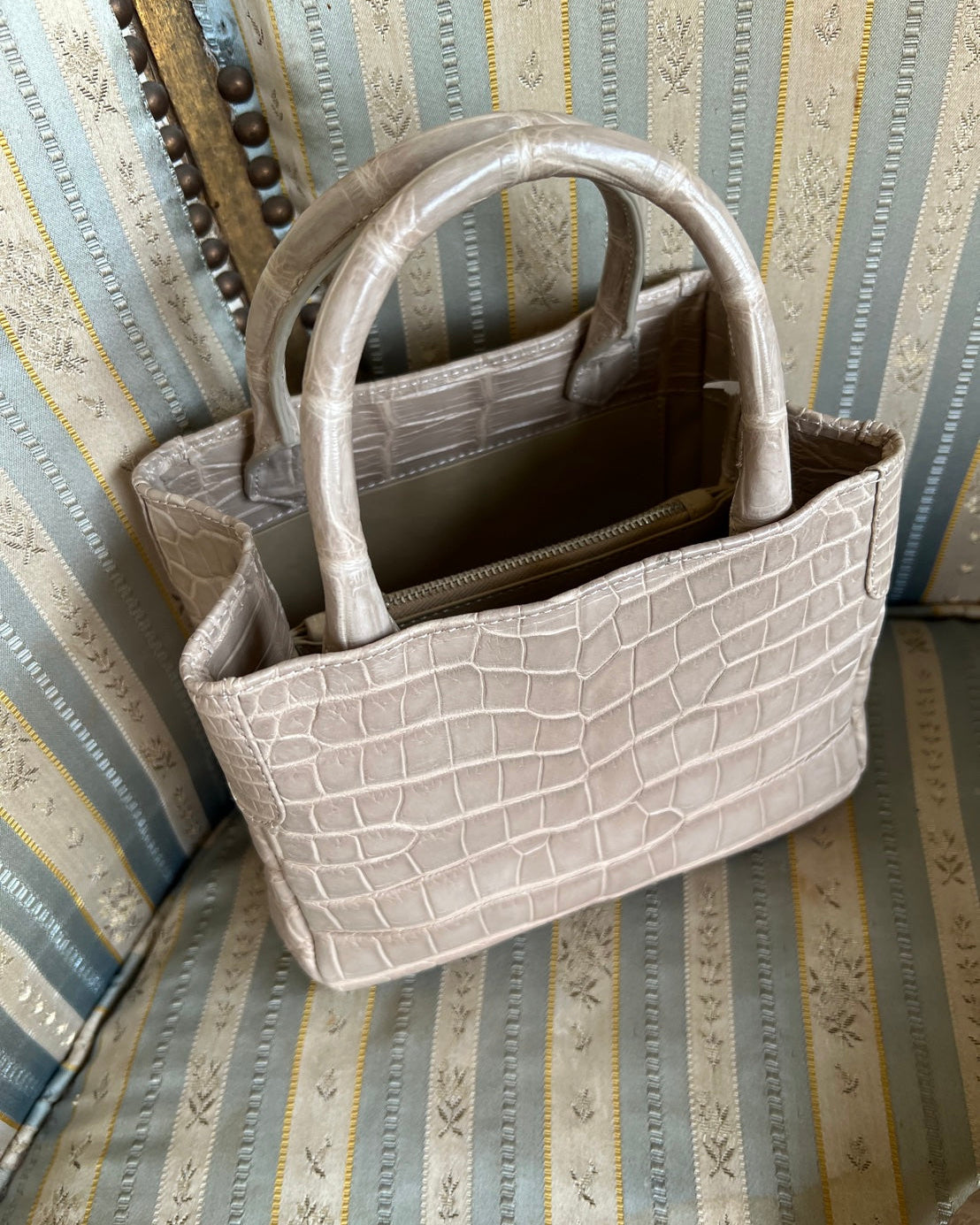 A structured handbag with a crocodile-embossed texture in a light taupe-beige color, resting on a striped fabric armchair.