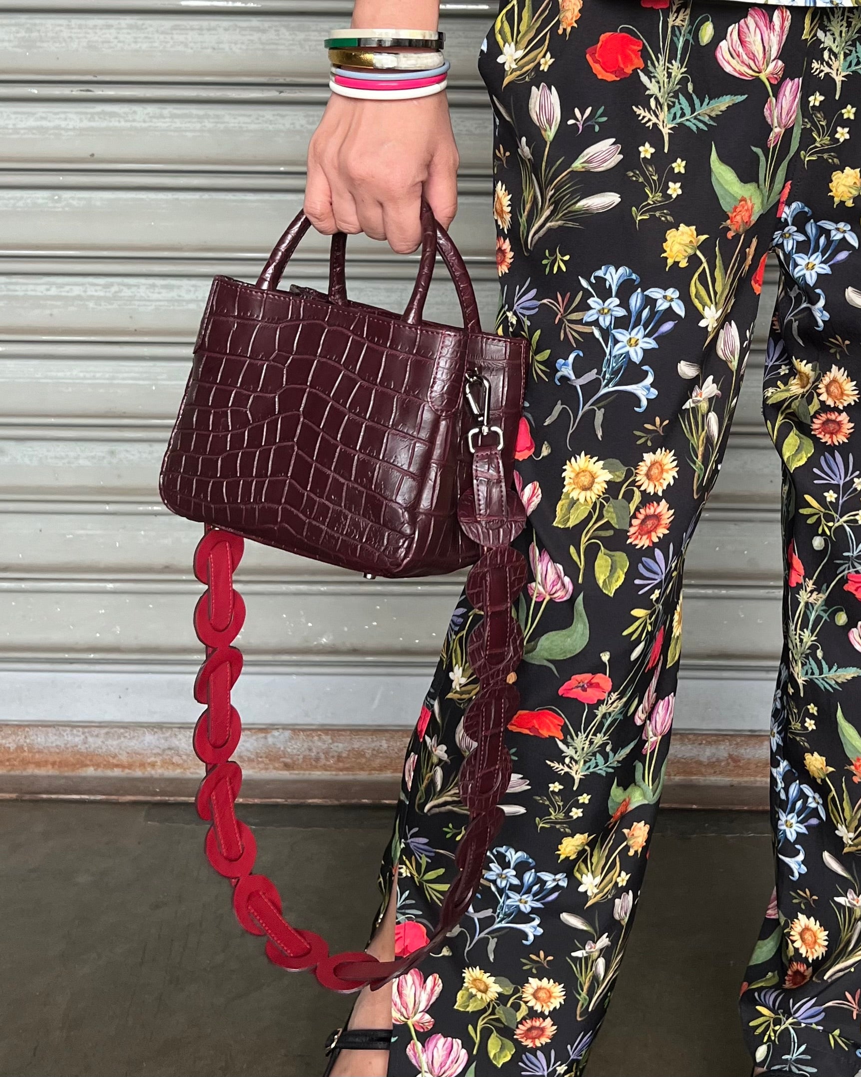Person holding a maroon handbag with a red strap, wearing floral-patterned pants.