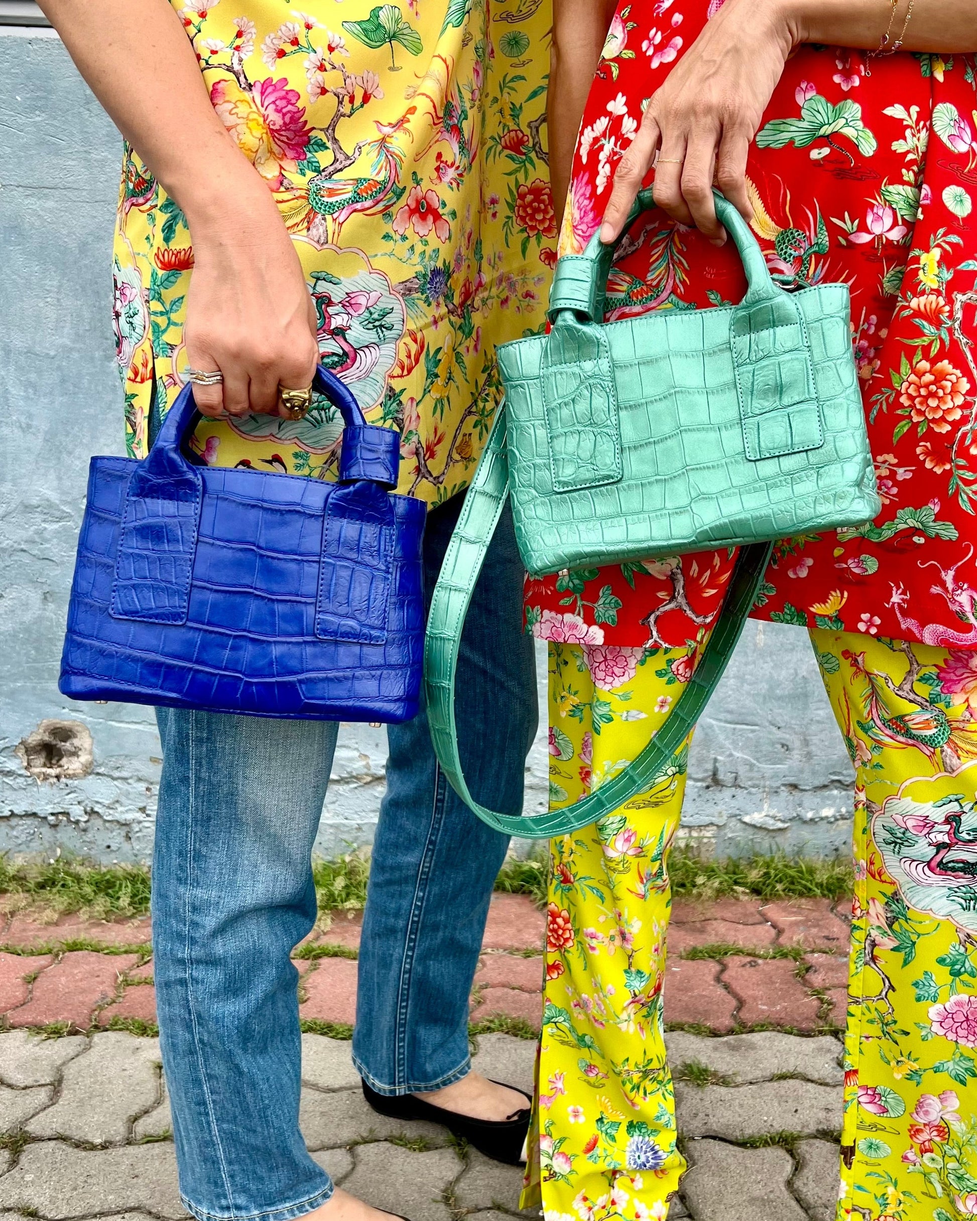 Two people holding colorful handbags against a gray wall.