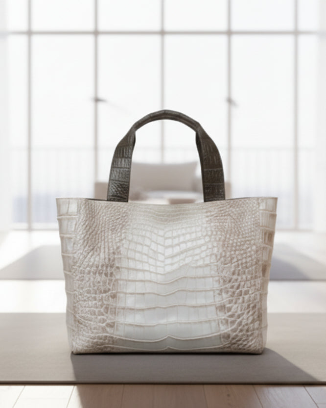 A white tote bag with crocodile-like texture, placed on a chair with a green leafy pattern. The bag has two handles and a zipper.