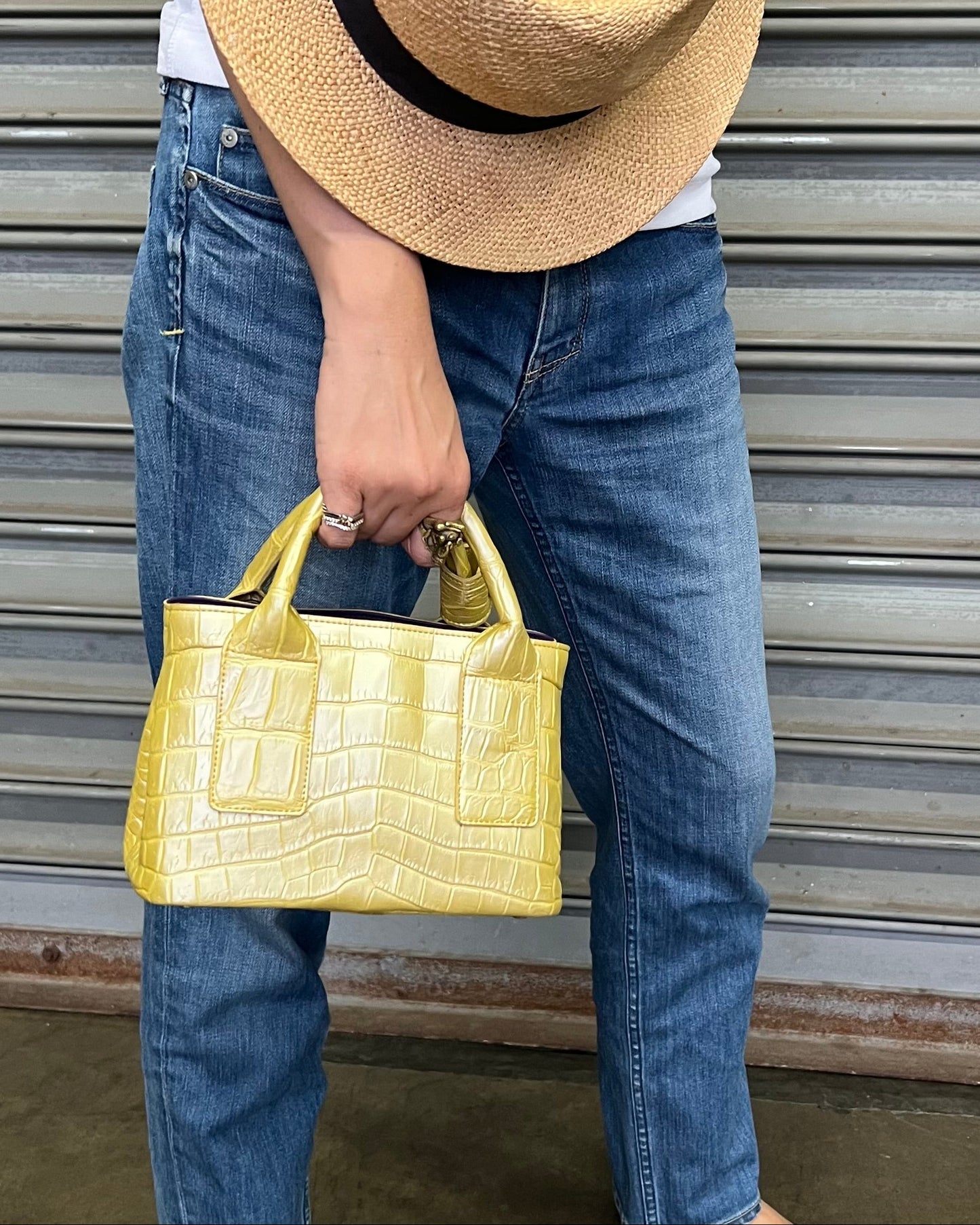 A person holding a golden yellow, crocodile leather handbag with a straw hat on.