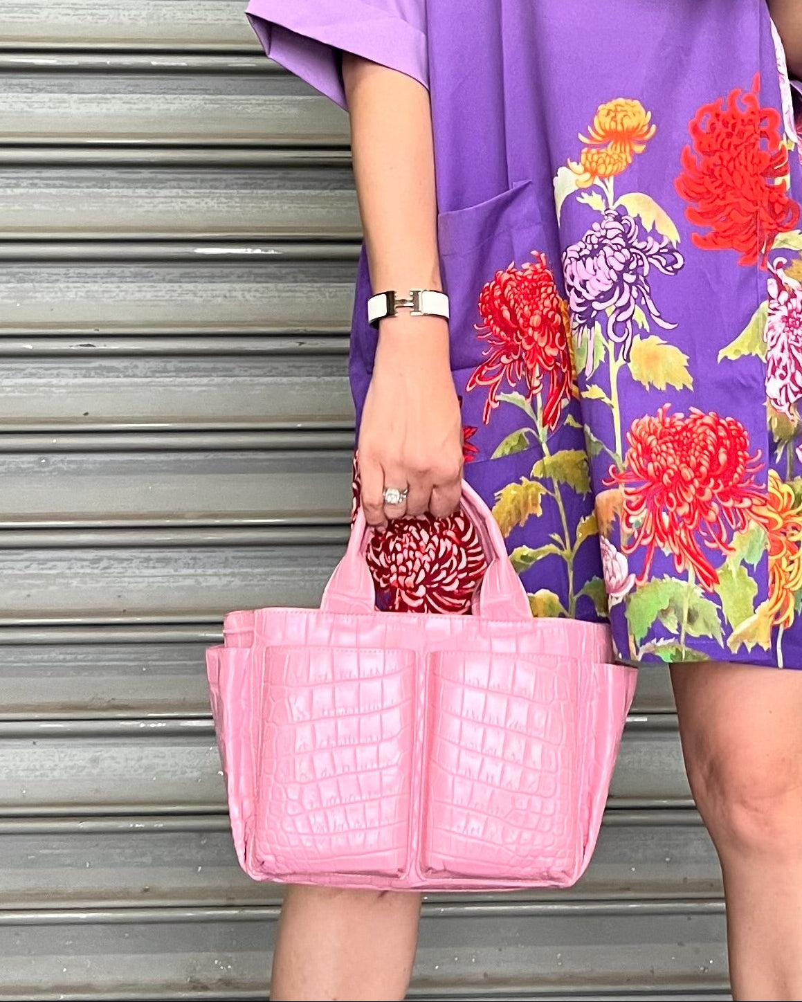 Person wearing a purple floral dress holding a pink bag against a metallic shutter background