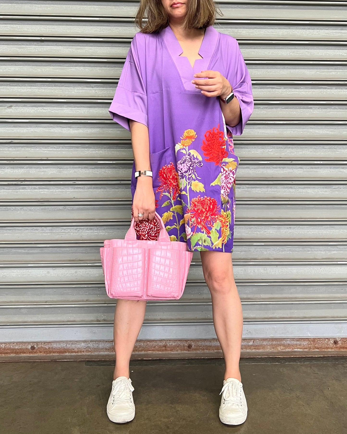 Person wearing a purple kimono-style outfit with floral patterns, holding a pink bag against a metallic shutter background.