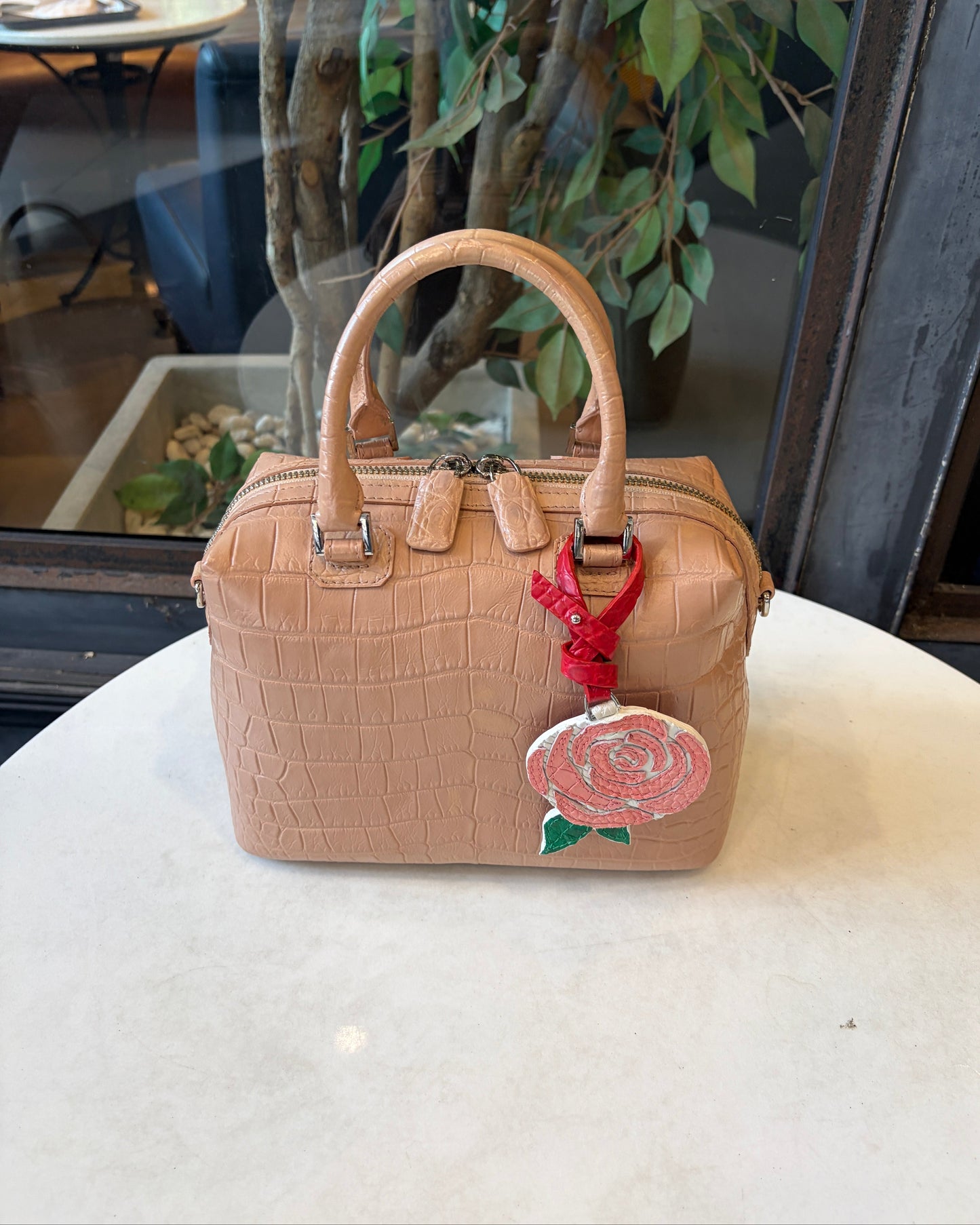 A structured tan crocodile-embossed handbag with dual top handles, a top zipper, and a red-and-white embroidered rose charm, displayed on a white tabletop with a cafe interior visible in the background.