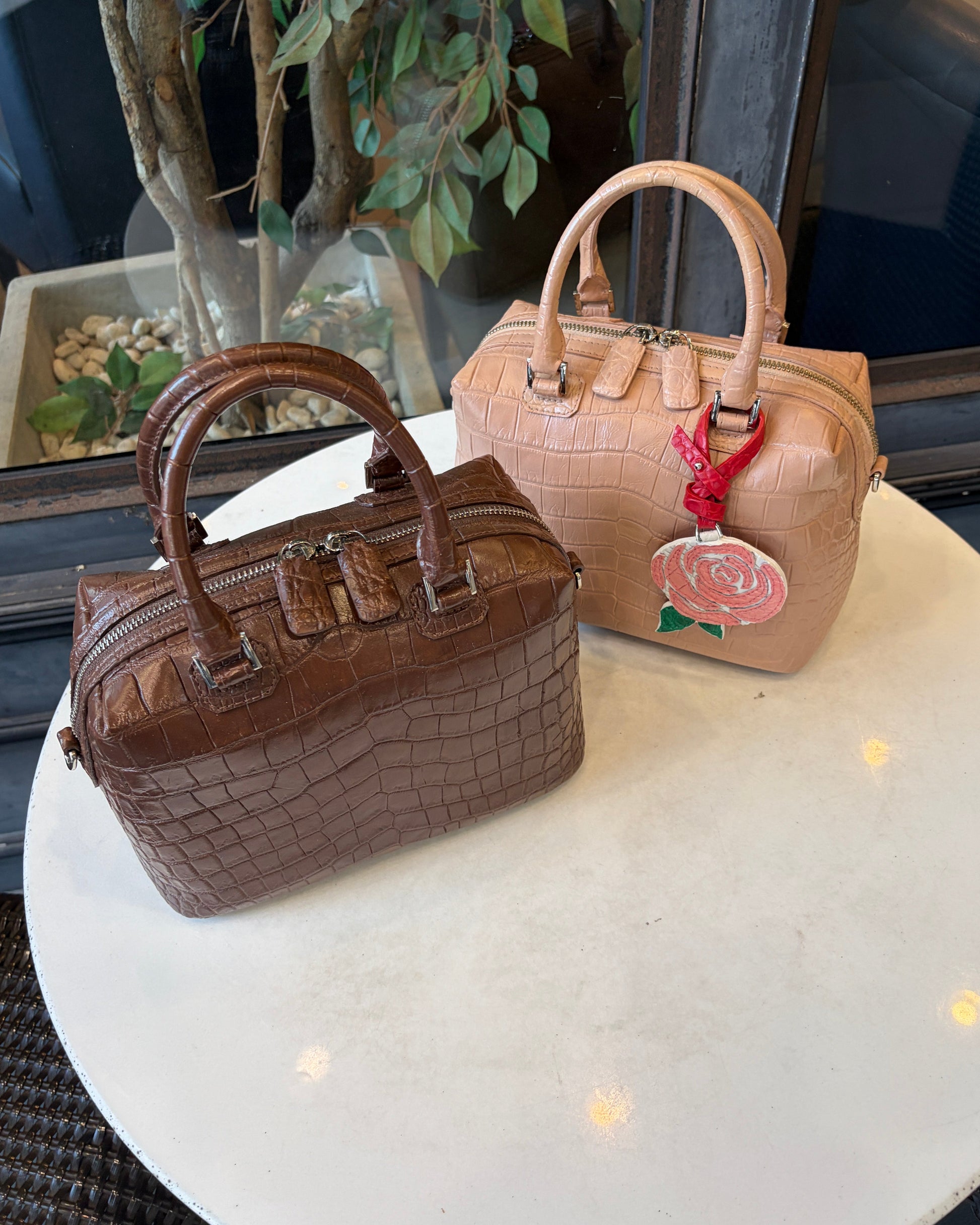 Two structured handbags displayed on a round white table in a boutique setting. The bag in the foreground is dark brown crocodile-embossed leather with double top handles and a zip-around closure.