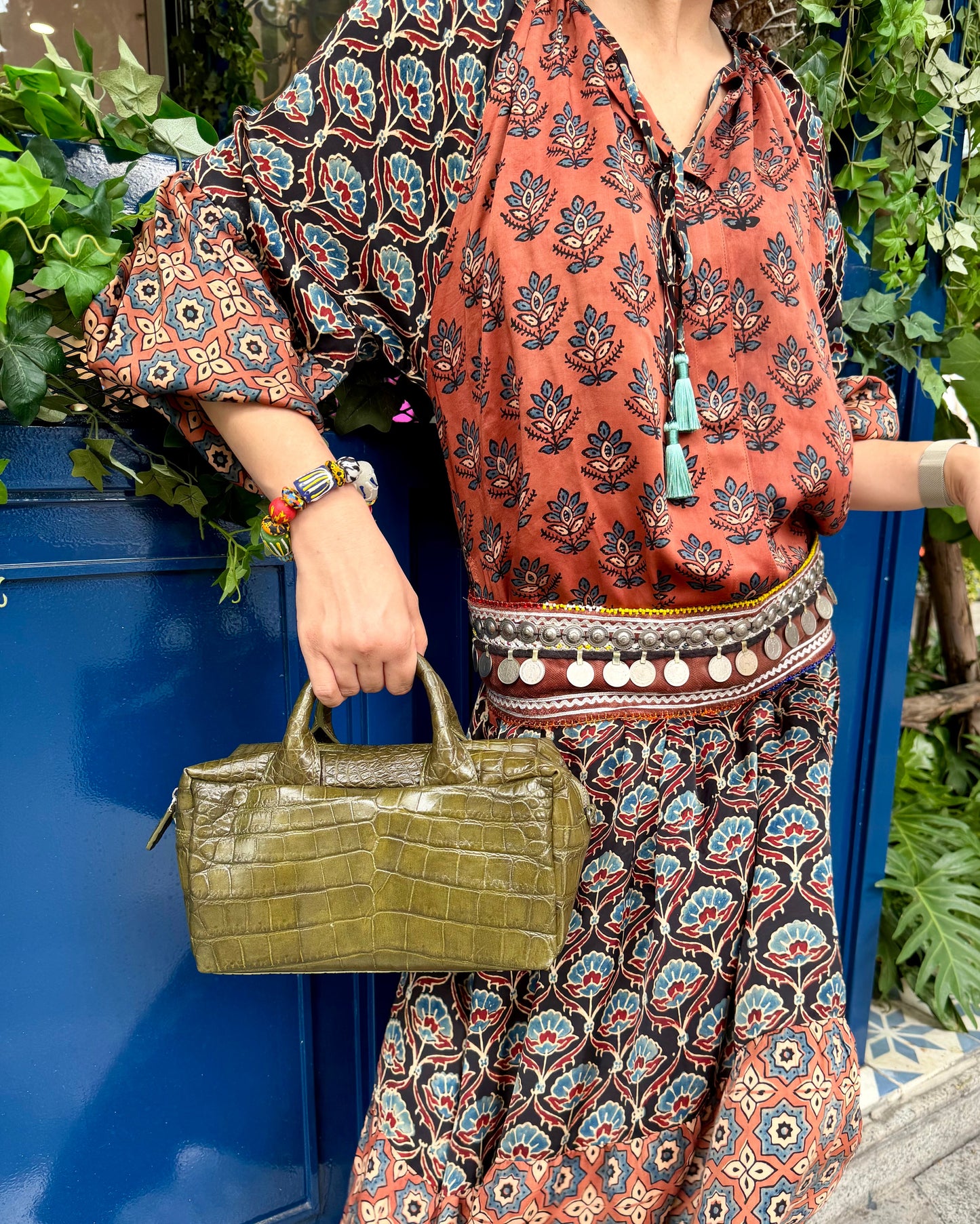 A woman wearing a bohemian-style rust orange and black printed blouse with tassel ties and a matching long skirt, holding a green crocodile-embossed handbag next to a blue door with green plants.