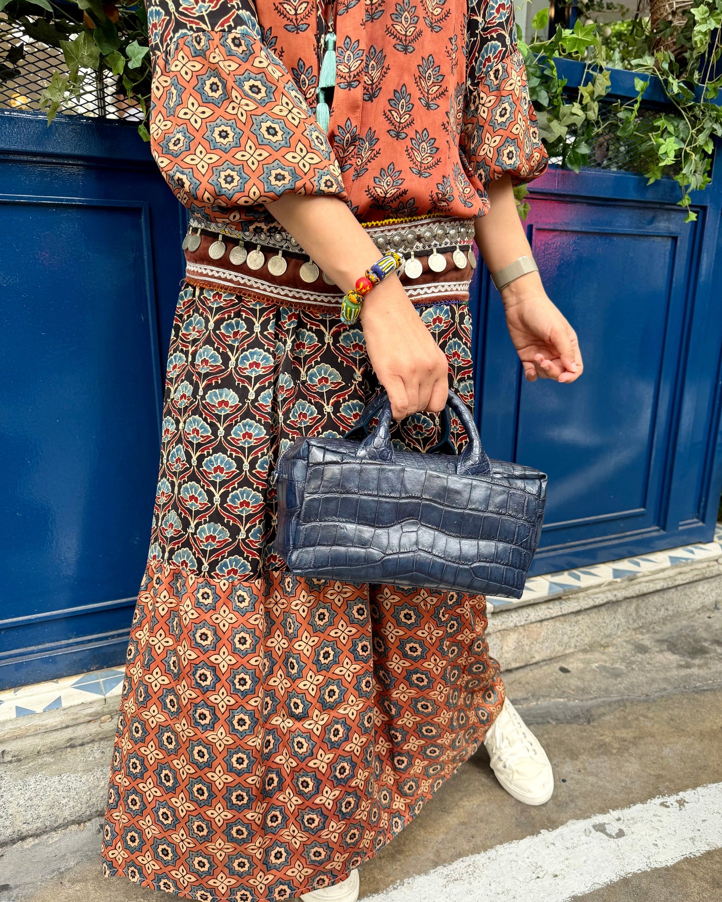 A person wearing a rust-orange and navy bohemian patterned maxi skirt and matching blouse, holding a dark blue crocodile-embossed handbag, standing in front of a blue wooden fence.
