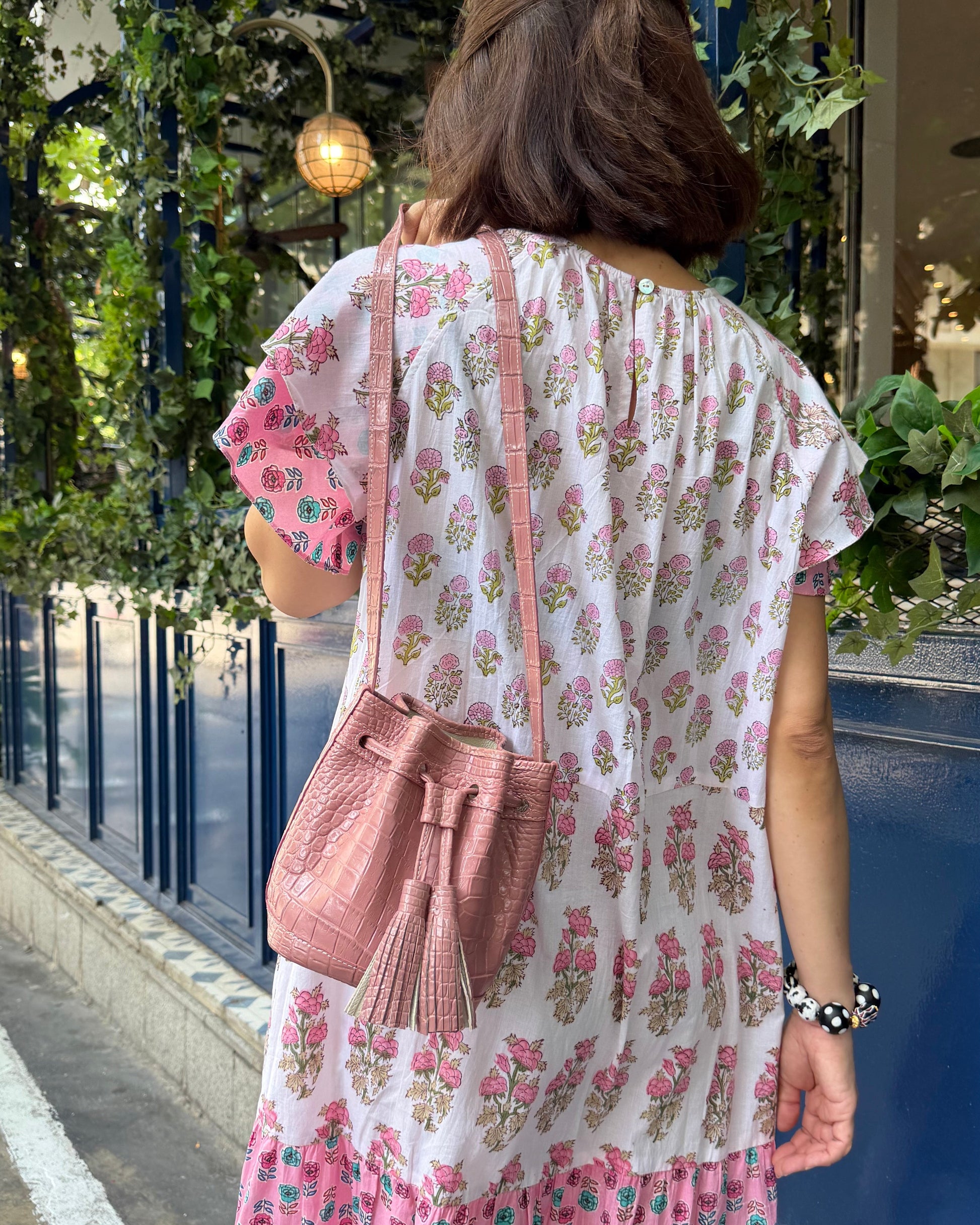A woman viewed from behind wearing a pink and white floral tiered dress and carrying a pink drawstring shoulder bag, standing on a sidewalk in front of a blue storefront decorated with green plants.
