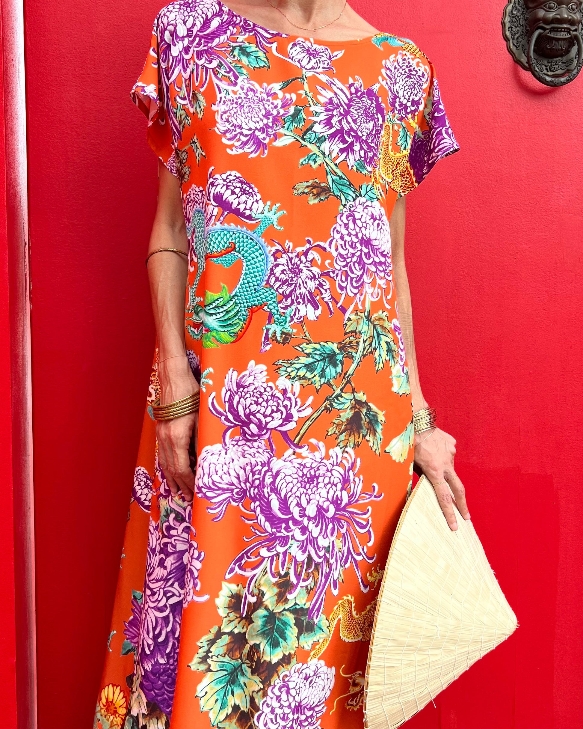 A woman wearing an orange maxi dress with a chrysanthemum and dragon pattern, featuring a boat-neckline and long sleeves.