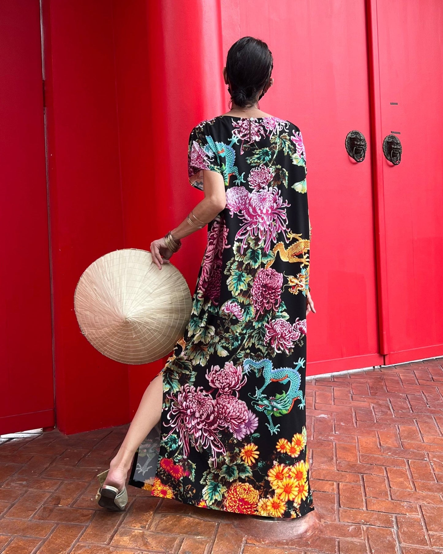 Person wearing a colorful floral dress holding a woven bag against a red wall.