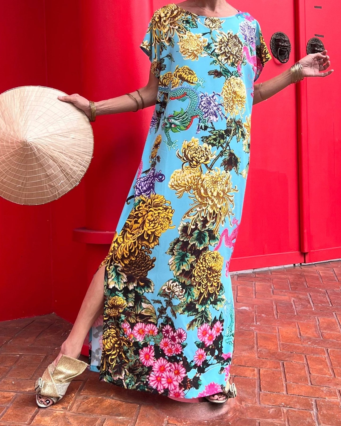Person wearing a colorful floral dress holding a conical hat against a red wall.