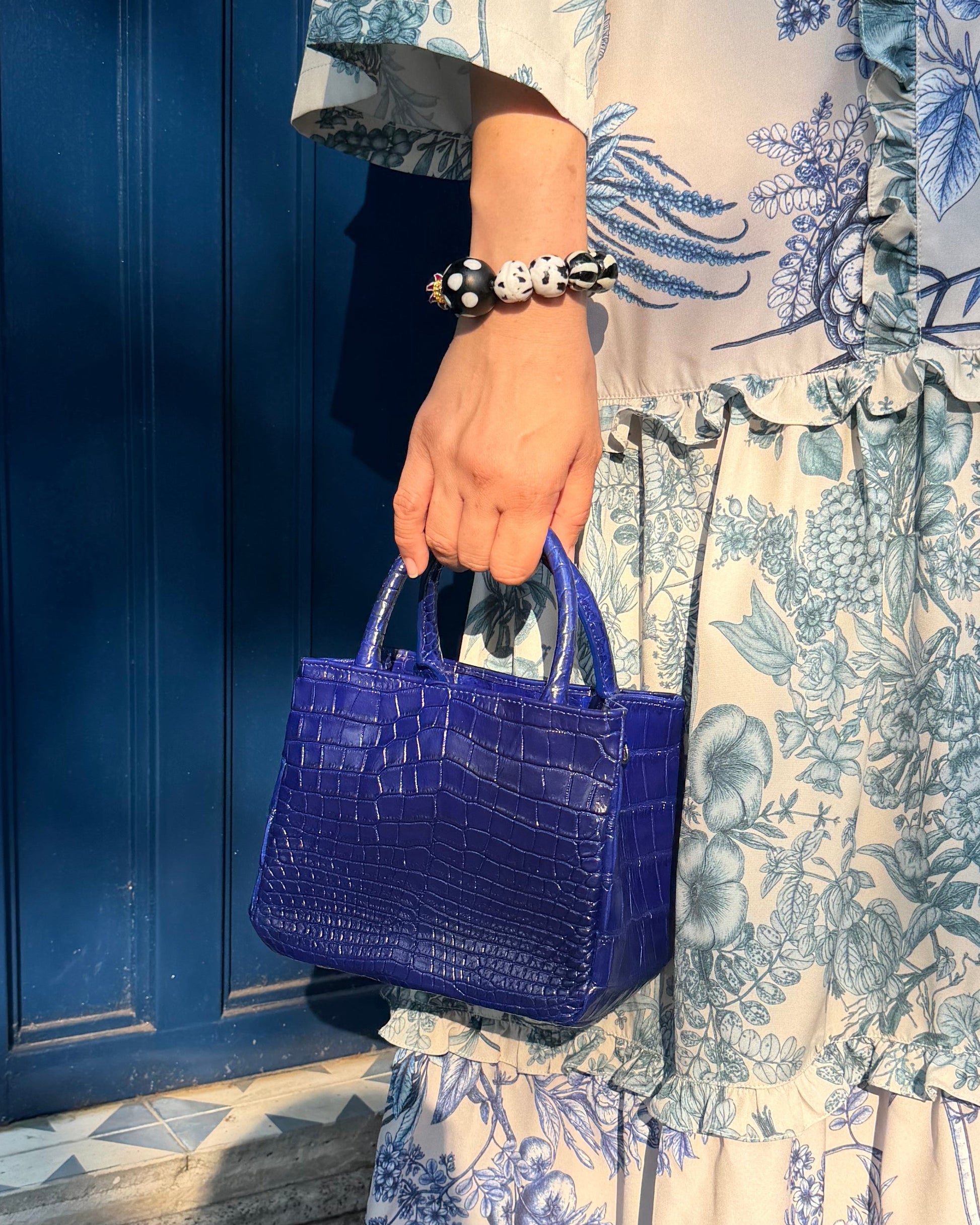 A person wearing a tiered cream dress with a blue floral toile pattern, holding a small textured purple handbag, standing outdoors next to a dark blue wooden fence.