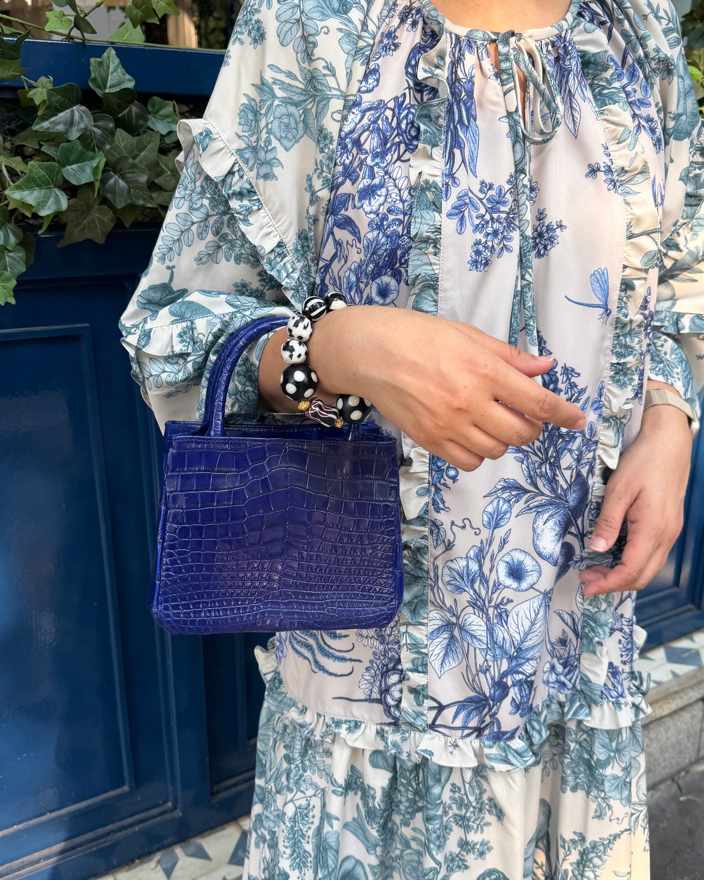 A person wearing a blue and white floral print dress or tunic with ruffled details, holding a structured cobalt blue crocodile-embossed handbag, standing in front of a dark blue door.