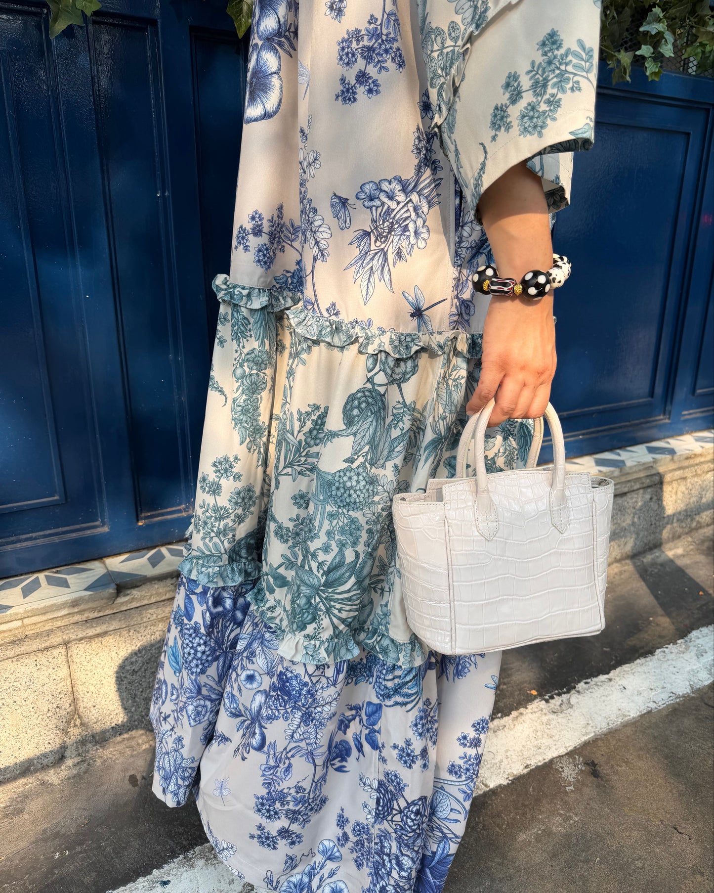 A person wearing a long cream dress with a blue floral toile pattern, holding a small white croc-embossed handbag, standing in front of a dark blue wooden door.