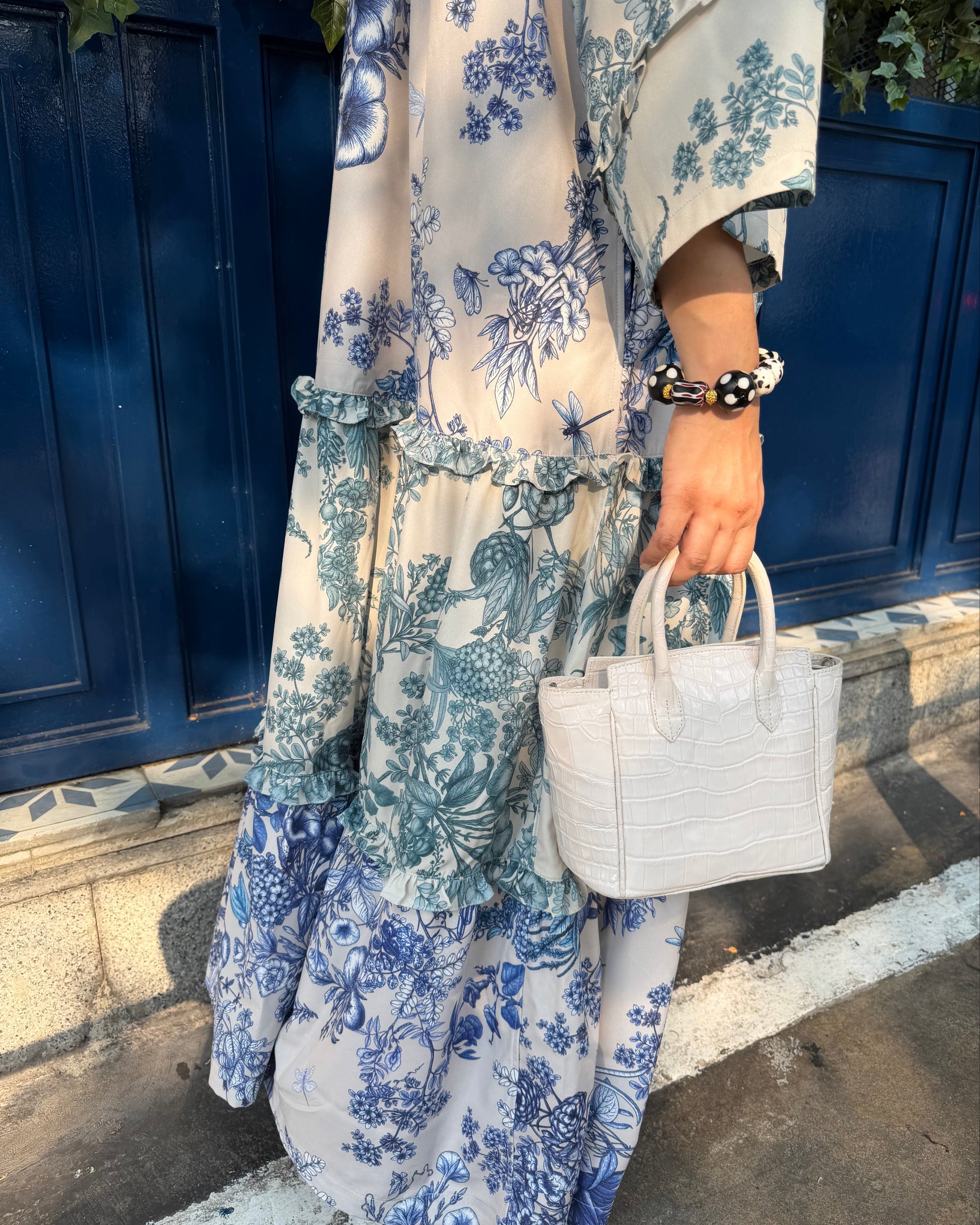 A person wearing a long cream dress with a blue floral toile pattern, holding a small white croc-embossed handbag, standing in front of a dark blue wooden door.