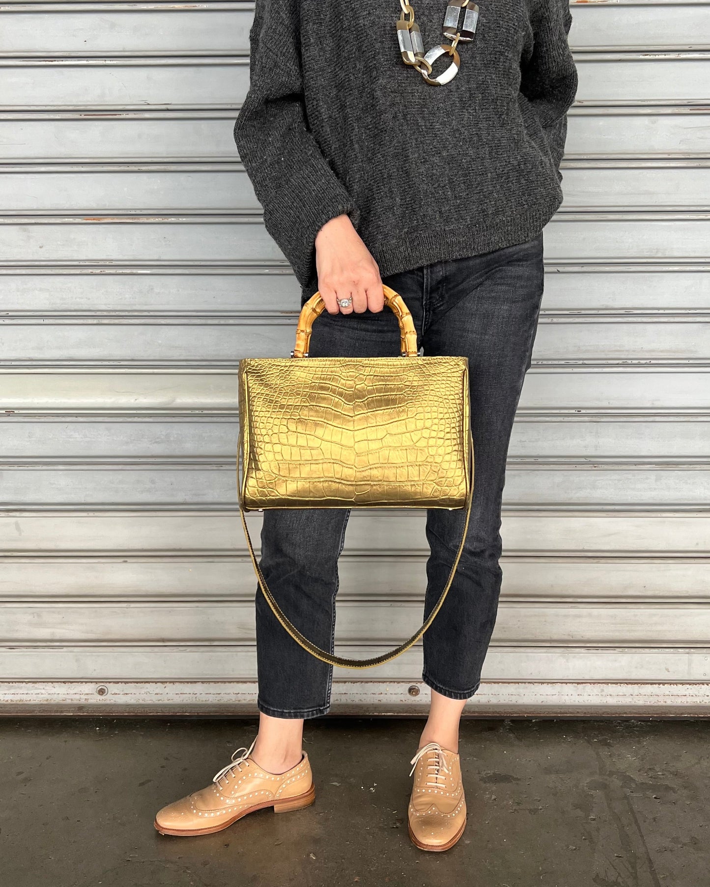 A person holding a gold-colored, crocodile leather handbag with a bamboo-style handle.