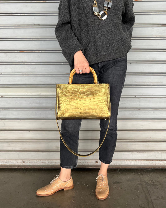 A person holding a gold-colored, crocodile leather handbag with a bamboo-style handle.