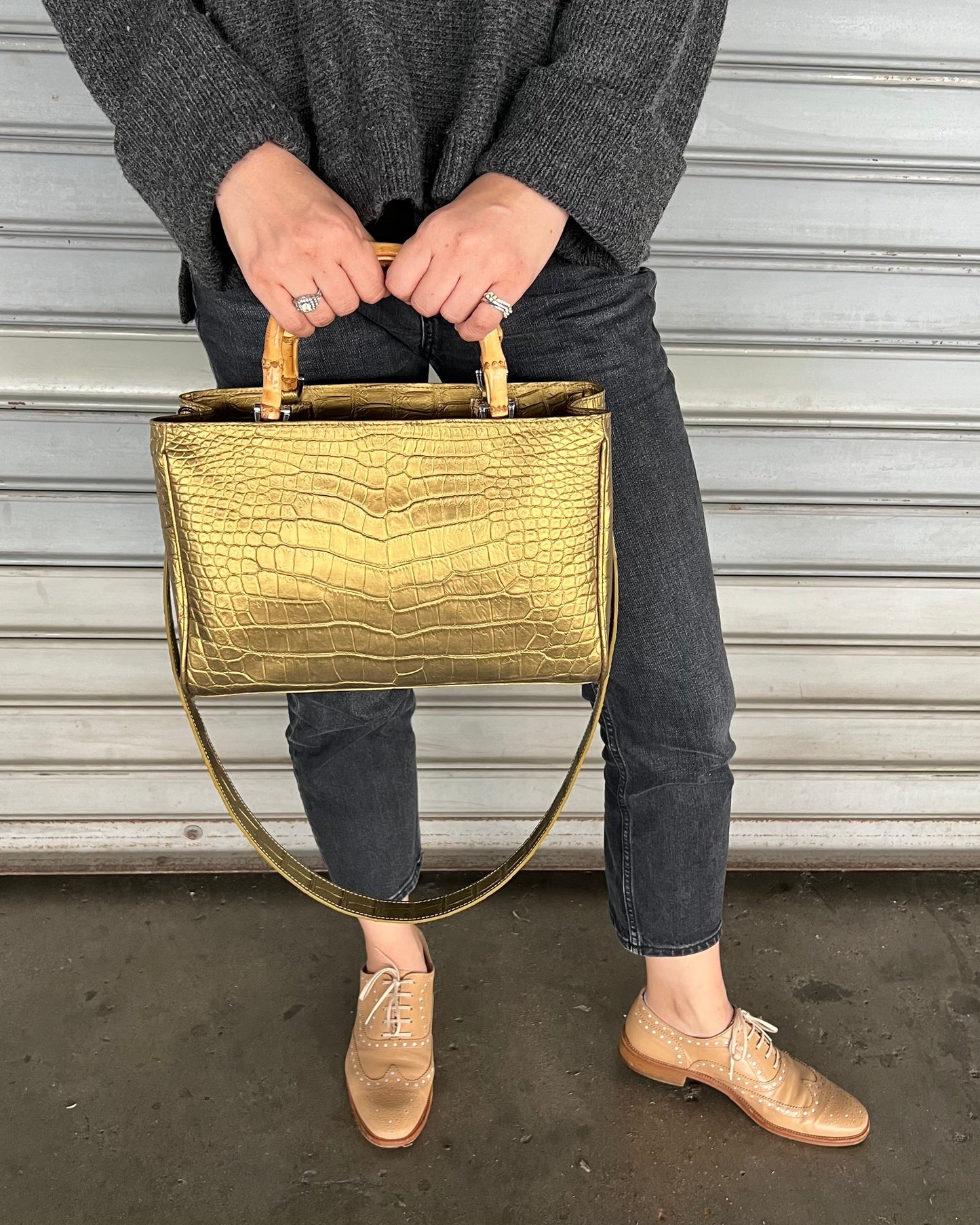 A person holding a gold-colored, crocodile leather top handle bag with a beige lining.