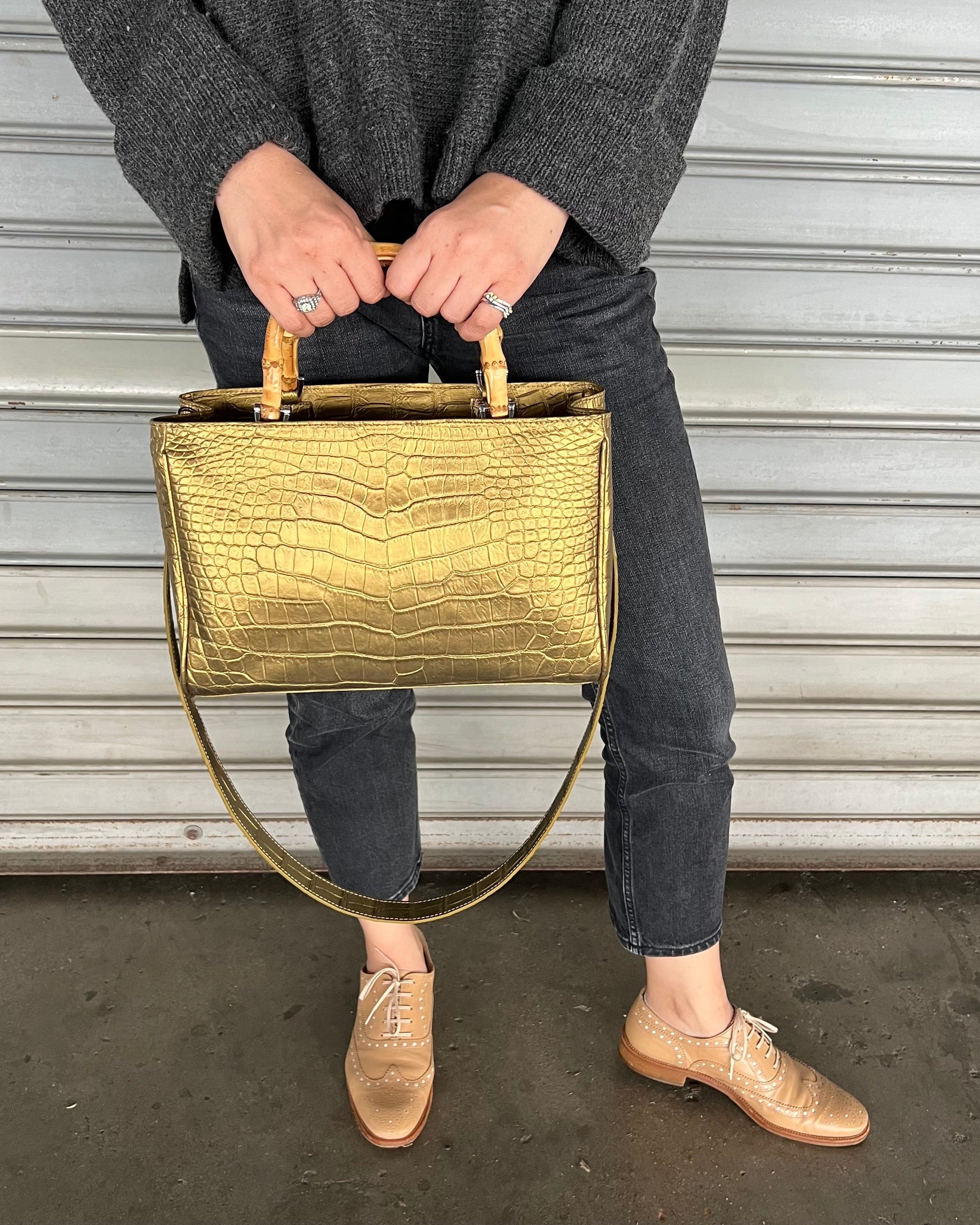 A person holding a gold-colored, crocodile leather top handle bag with a beige lining.