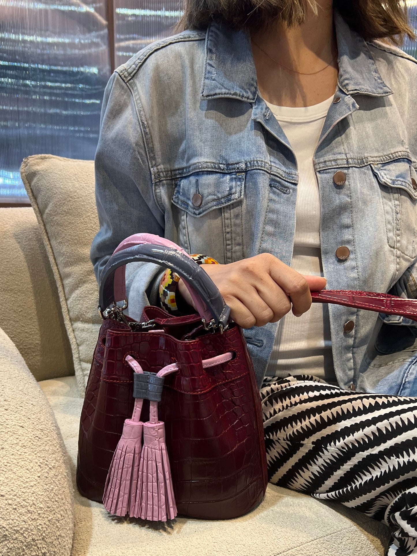 Person holding a maroon handbag with tassels on a beige couch.