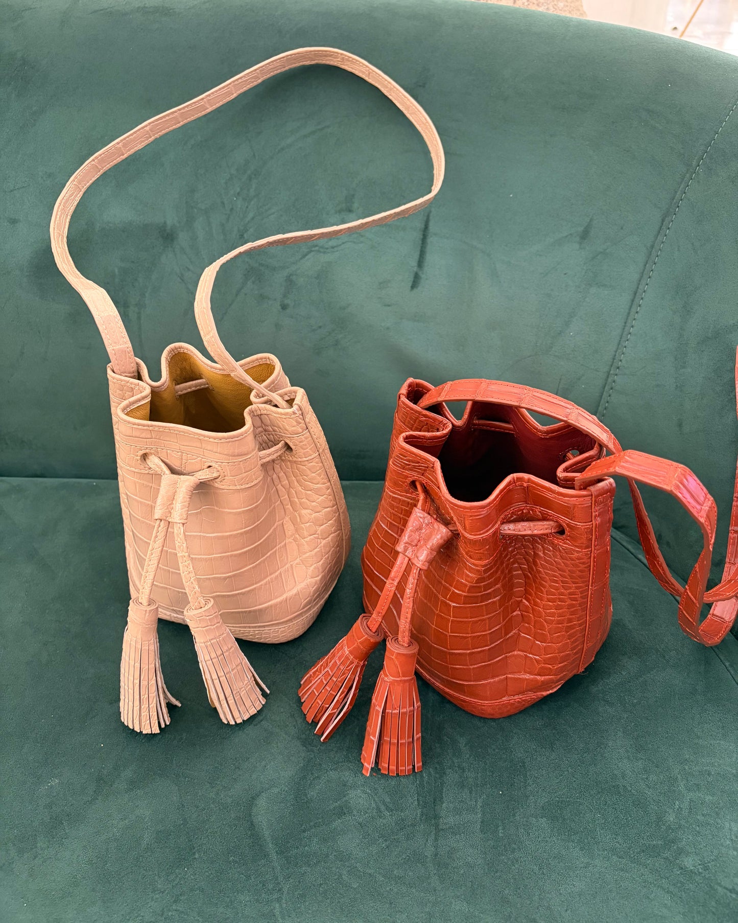 Two crocodile-embossed leather bucket bags with tassel drawstrings, one in light beige and one in terracotta orange, displayed on a green velvet sofa.