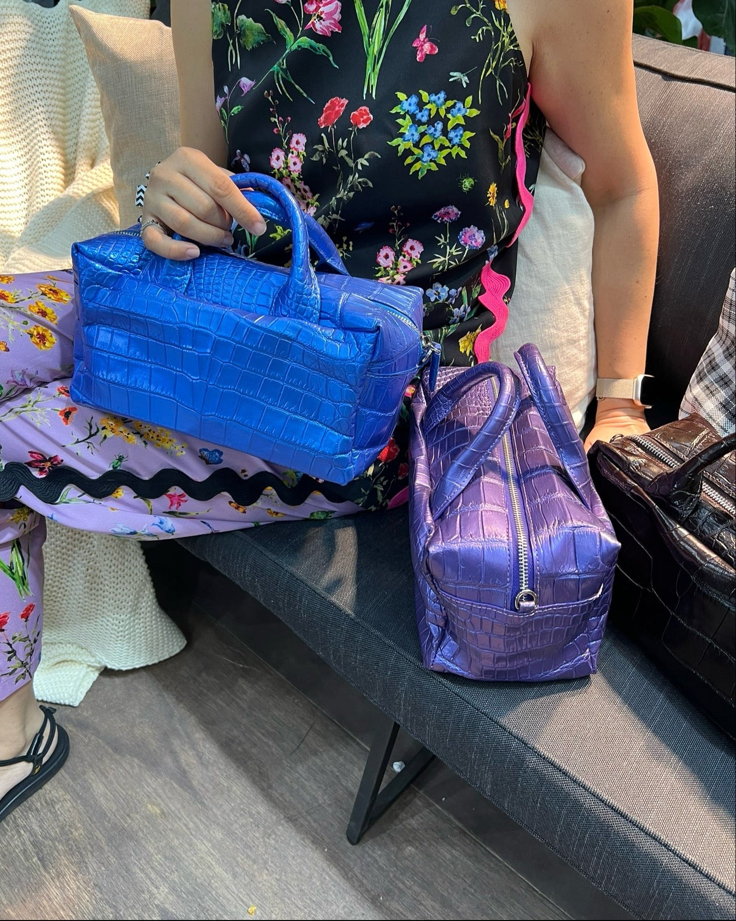 A person seated on a sofa holds a bright blue crocodile-embossed handbag, with a purple crocodile-embossed handbag resting beside them and a black handbag partially visible on the right.