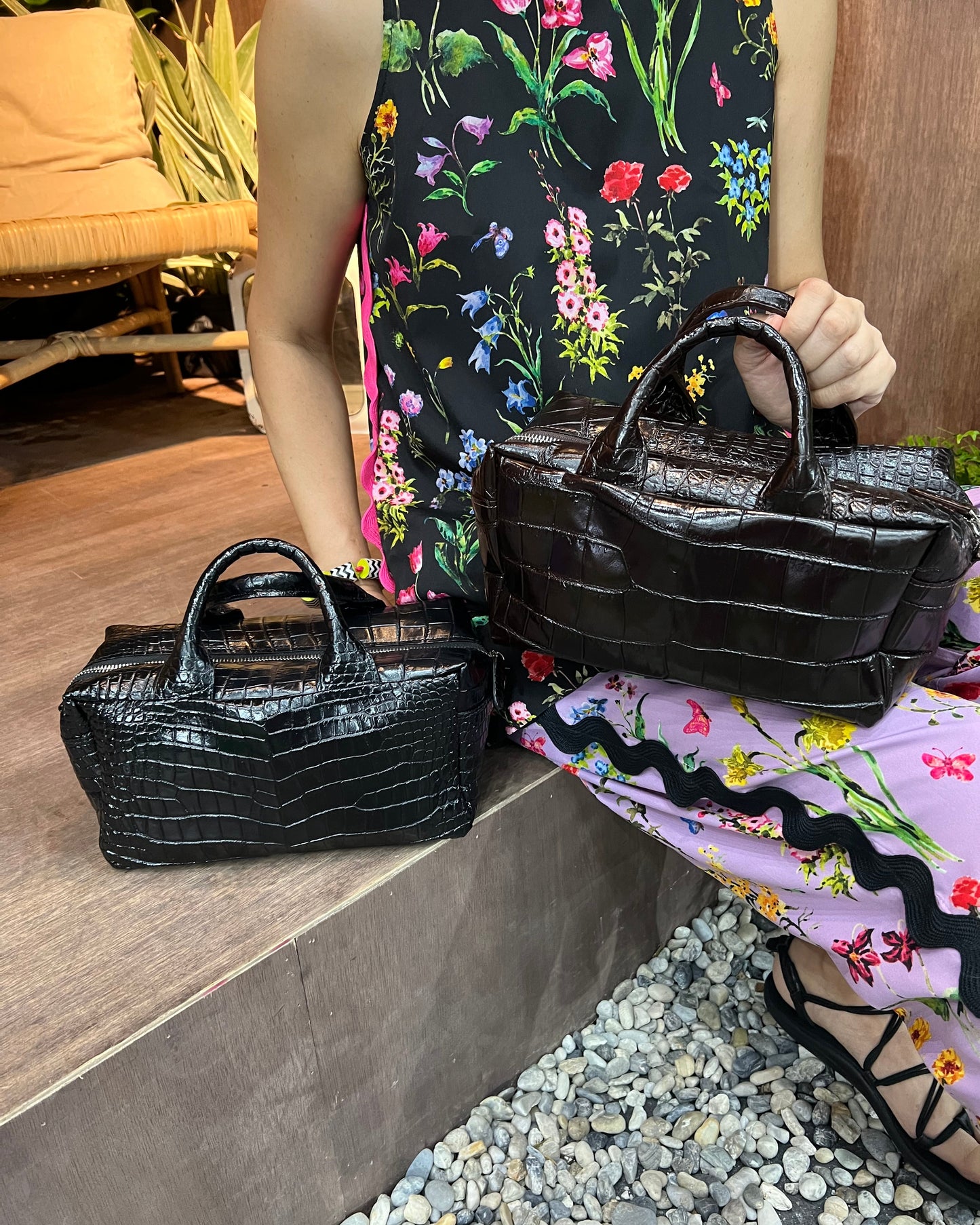 Person holding two black handbags with floral-patterned pants in the background