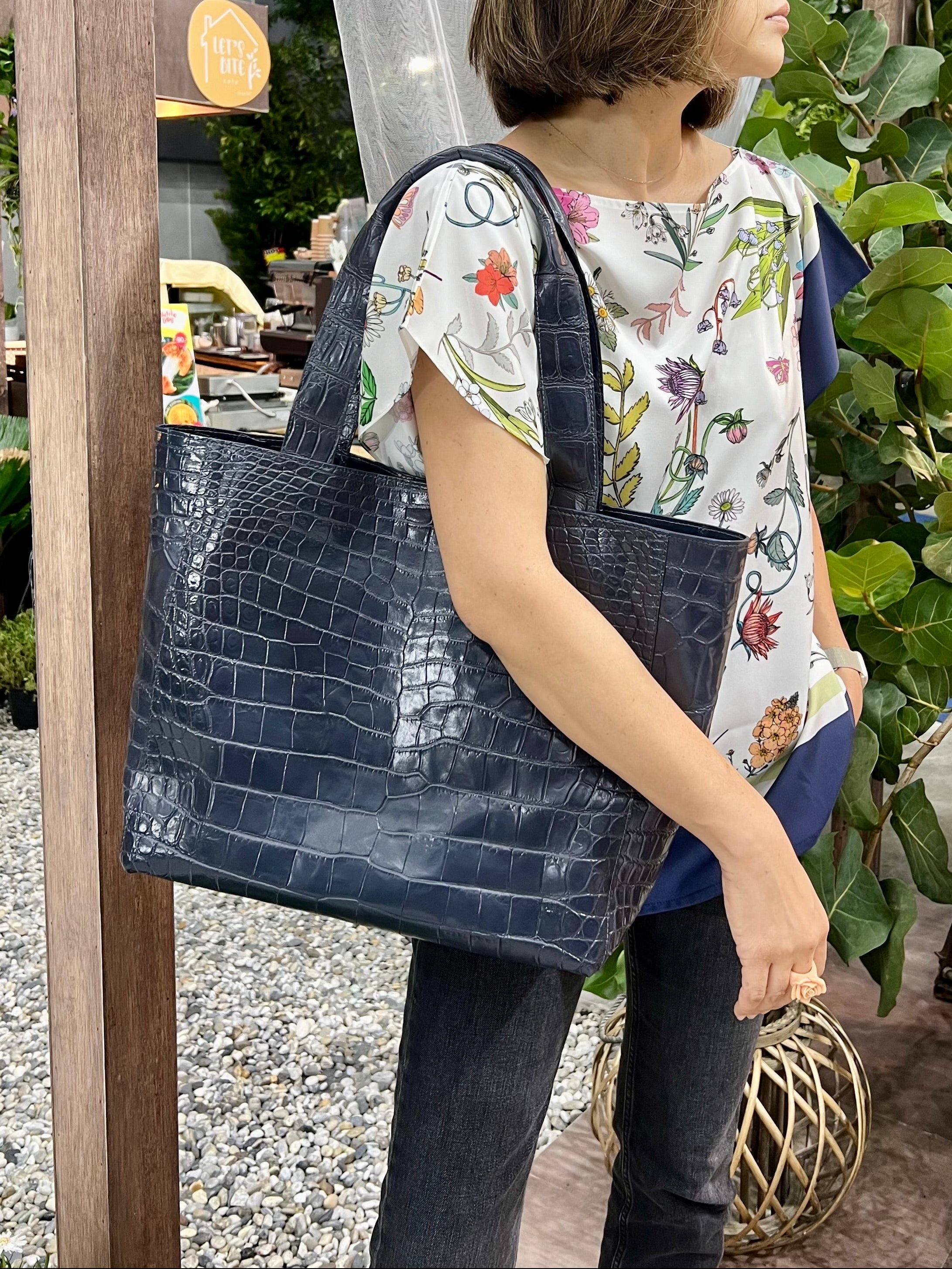 Woman holding a navy blue handbag in an outdoor setting with plants and wooden structures.