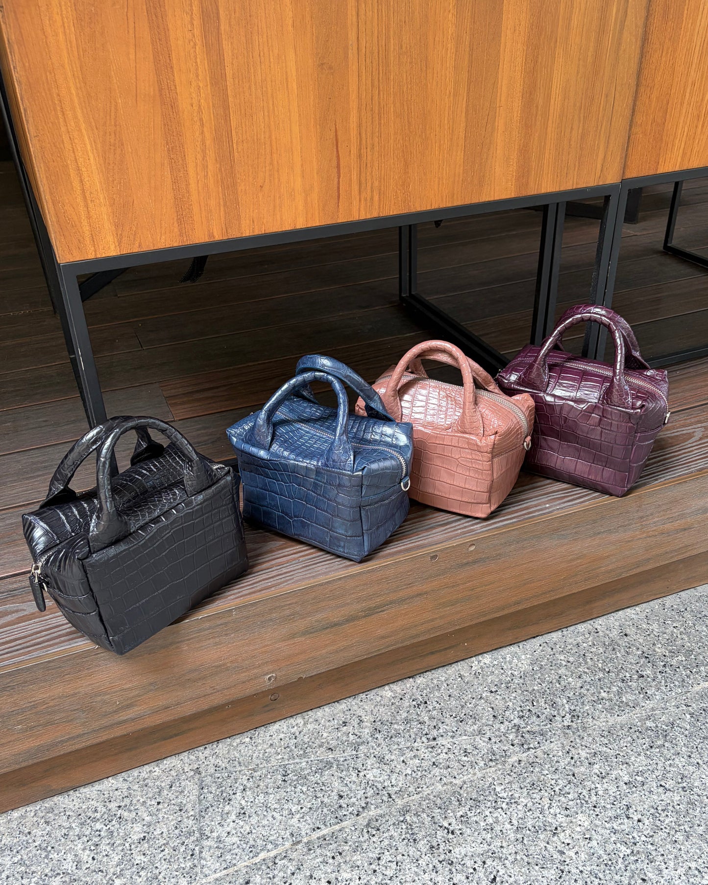 Four small structured handbags with crocodile-embossed leather are lined up on a wooden platform in front of a wooden cabinet. The bags are shown in black, blue, dusty pink, and plum purple colors.