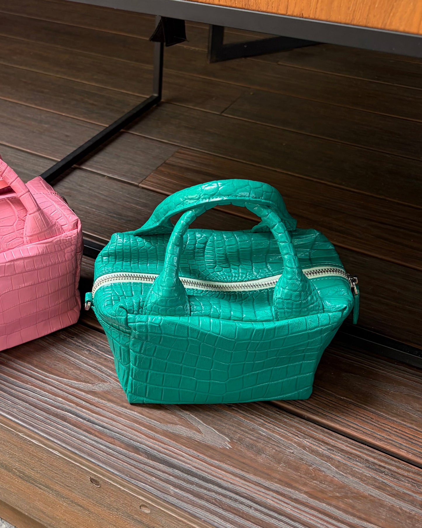 A small structured handbag with a crocodile-embossed texture in turquoise green, featuring dual top handles and silver zippers, resting on a dark wood floor next to a pink bag.