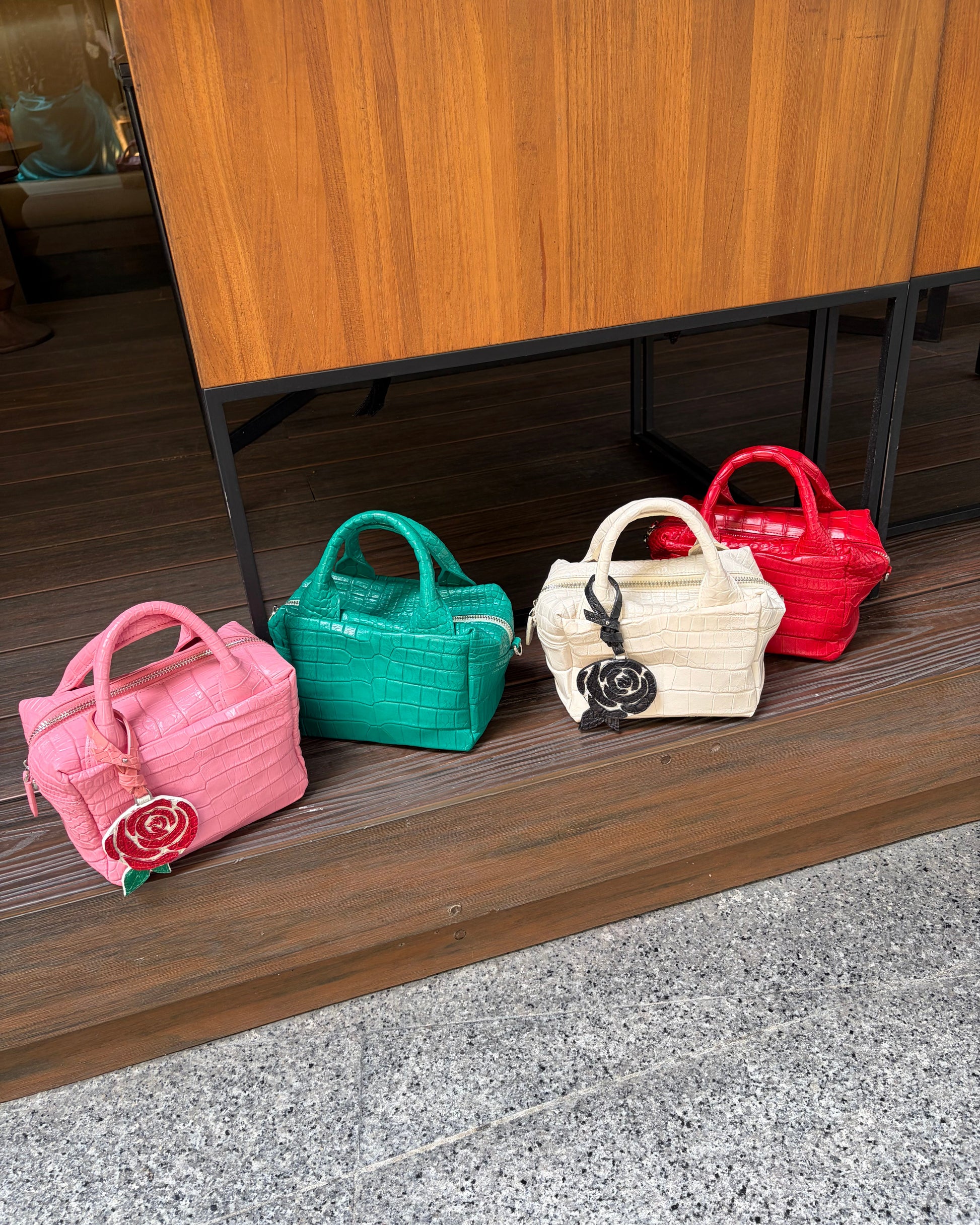 Four small woven handbags in pink, green, cream, and red are lined up on a wooden bench in front of a wooden cabinet.