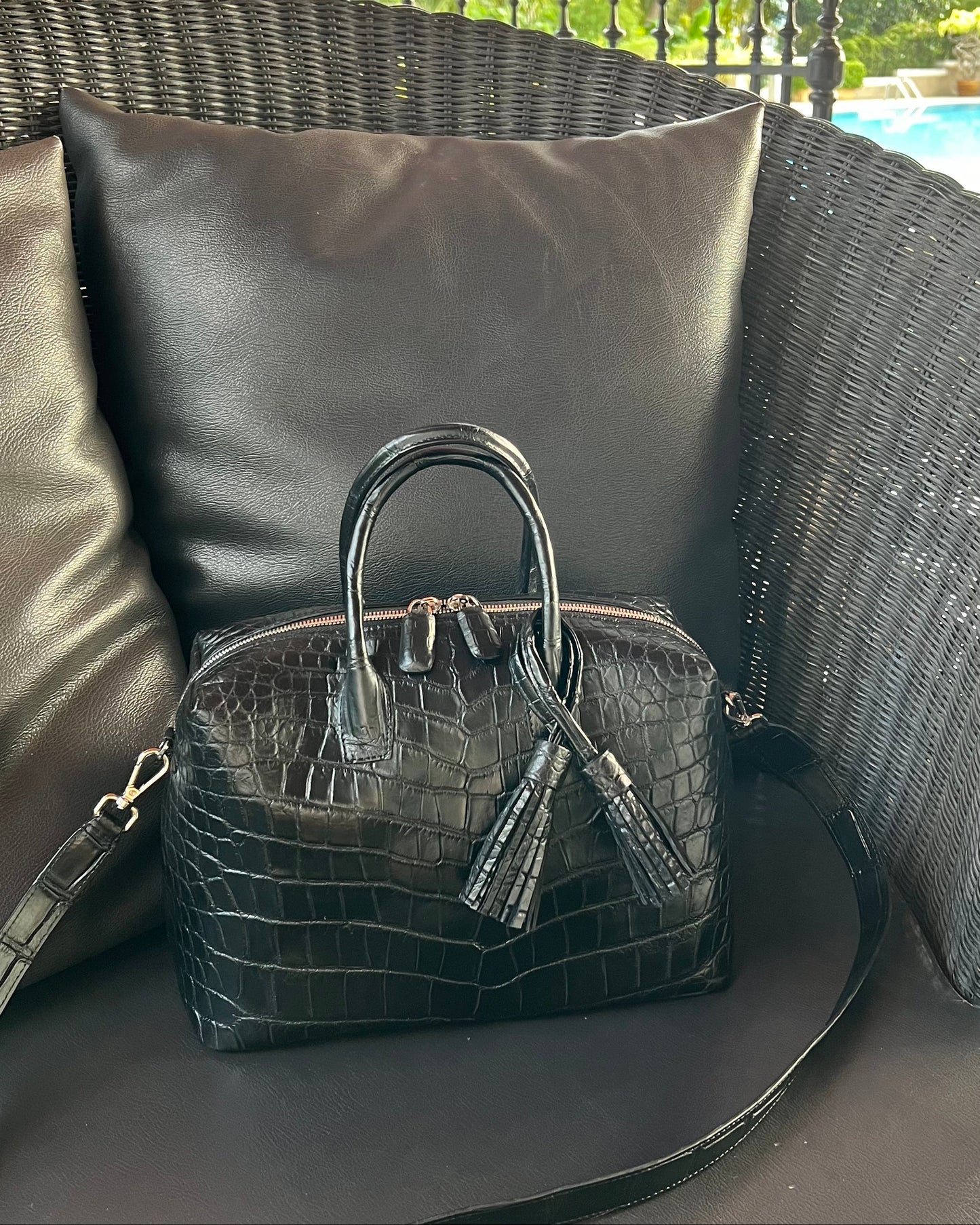 Black crocodile leather top handle bag with a flat, structured silhouette, placed on a black cushioned surface with a blurred background.