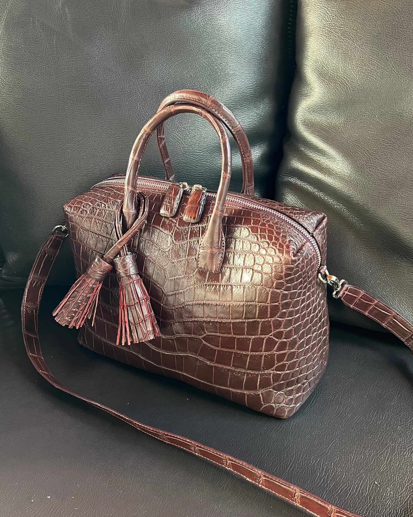 A brown crocodile leather top handle bag with black lining, displayed on a couch.