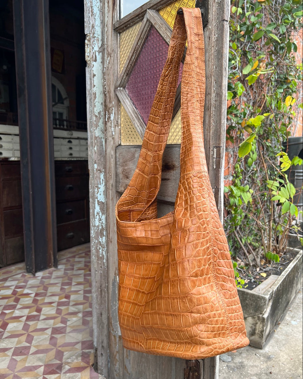 Brown textured handbag hanging on a wooden door with a patterned floor and plants in the background.