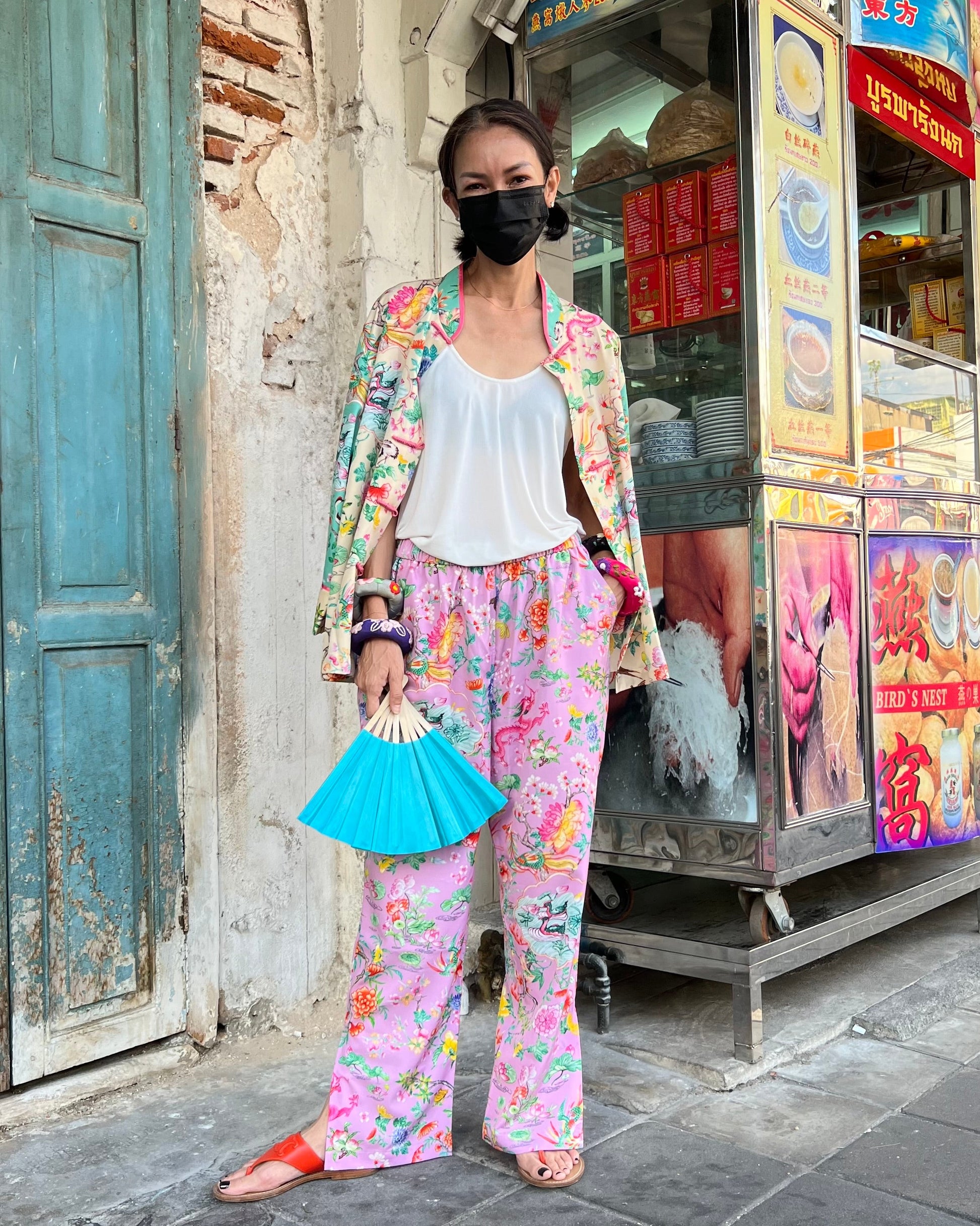 Woman in colorful outfit holding a blue fan standing in front of a traditional food stall.