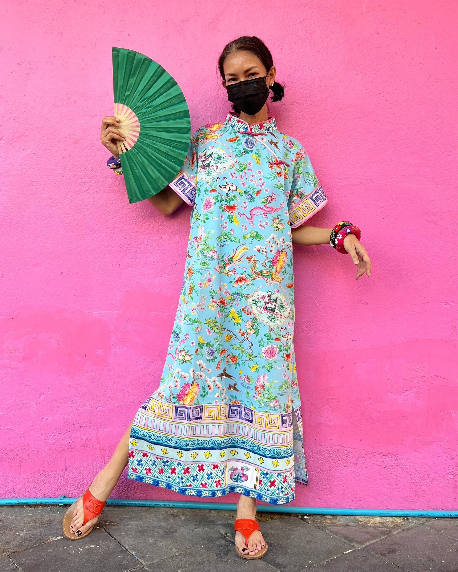 A woman posing in a blue traditional Chinese cheongsam dress with a mandarin collar and pankou knots closure, against a pink background.