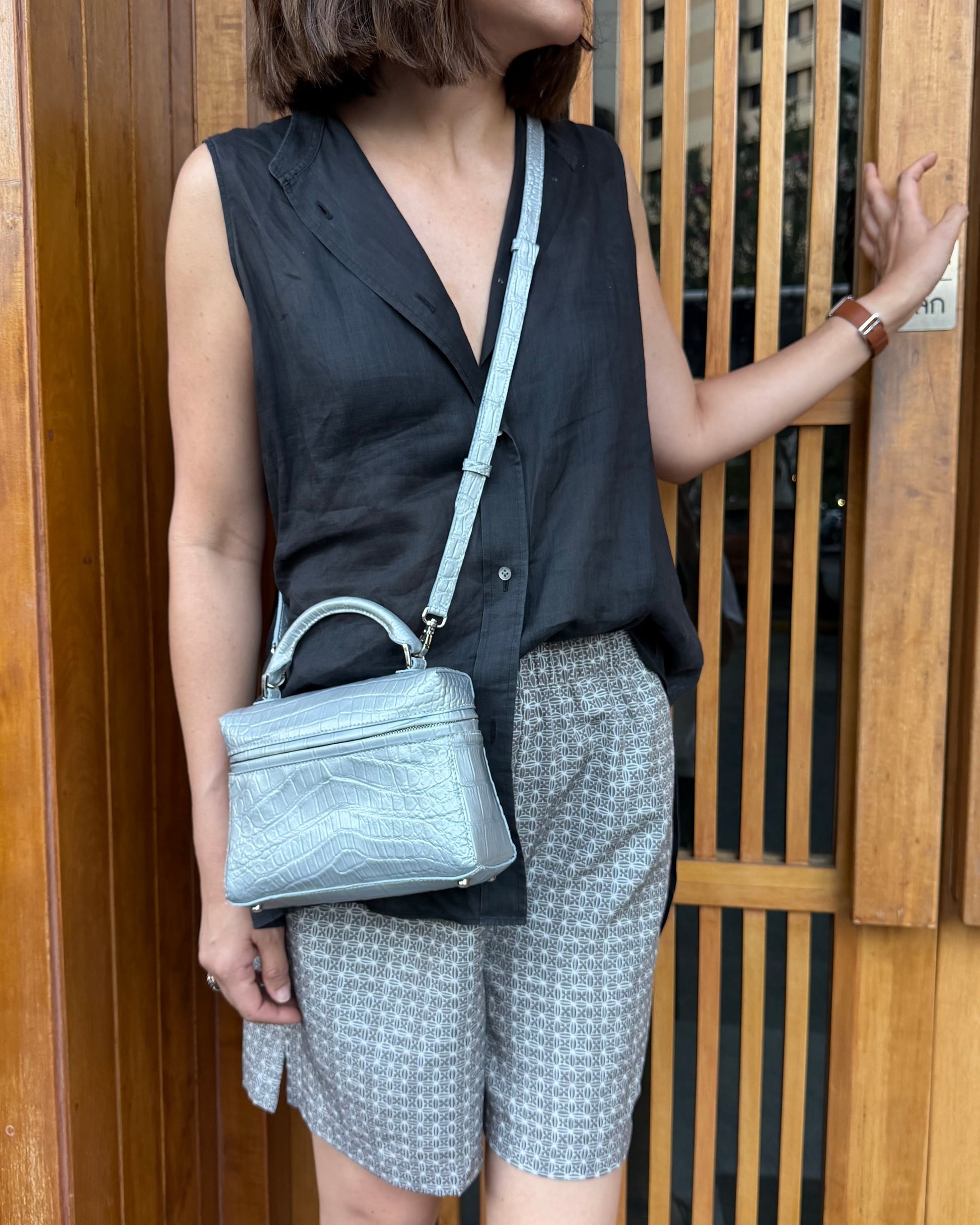 Person wearing a black sleeveless top and patterned shorts, holding a light blue handbag, standing in front of a wooden door.