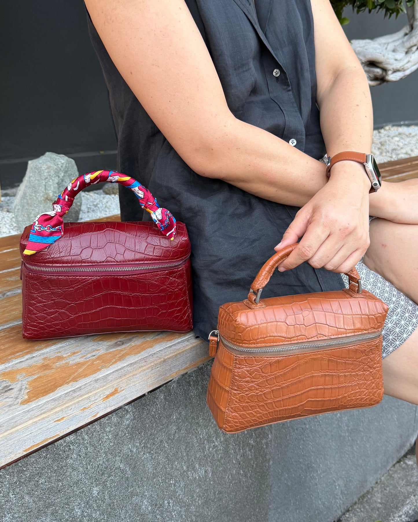A woman sitting on a wooden bench holds a small crocodile-embossed tan handbag with a top handle, while another matching red handbag rests beside her. The setting is outdoors on a bench with a dark wall and plant in the background.