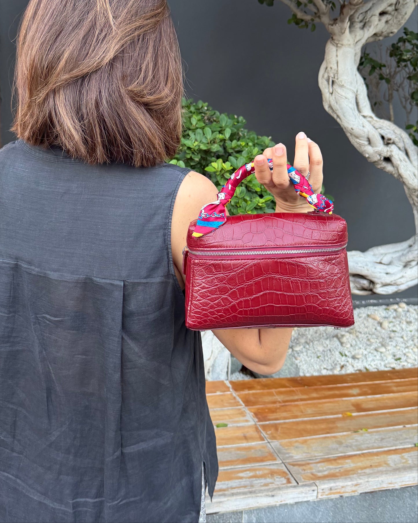 A woman viewed from behind holding a red crocodile-embossed handbag with a colorful patterned top handle in an outdoor garden setting.