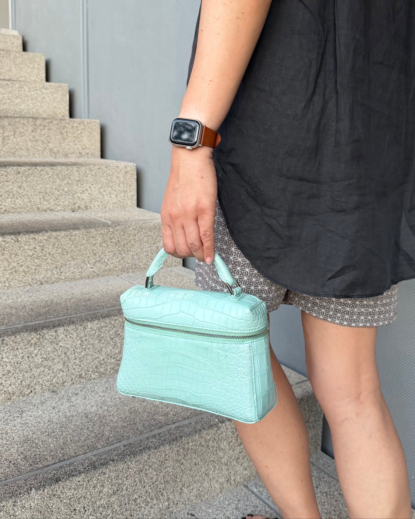 A person wearing a black dress and patterned shorts carries a small mint green crocodile-embossed handbag while walking down concrete stairs.