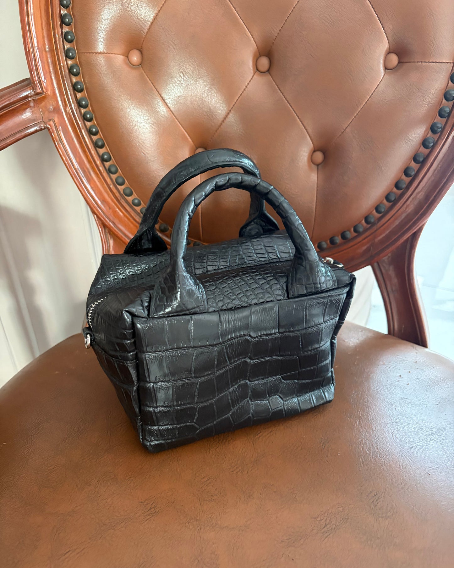 A small black crocodile-embossed leather handbag with double top handles, resting on a brown leather chair.