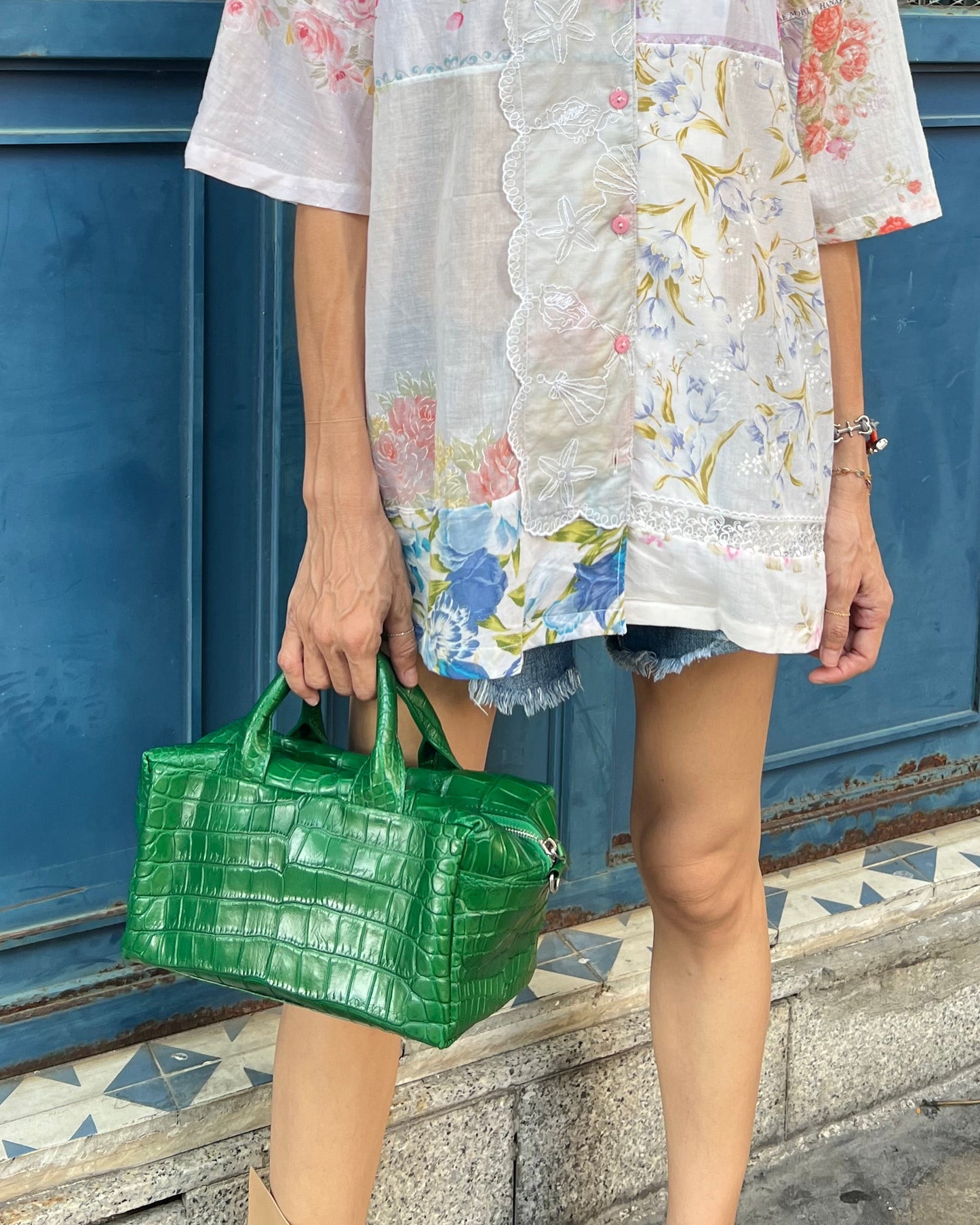 Woman in a floral dress holding a green handbag in front of a blue door.