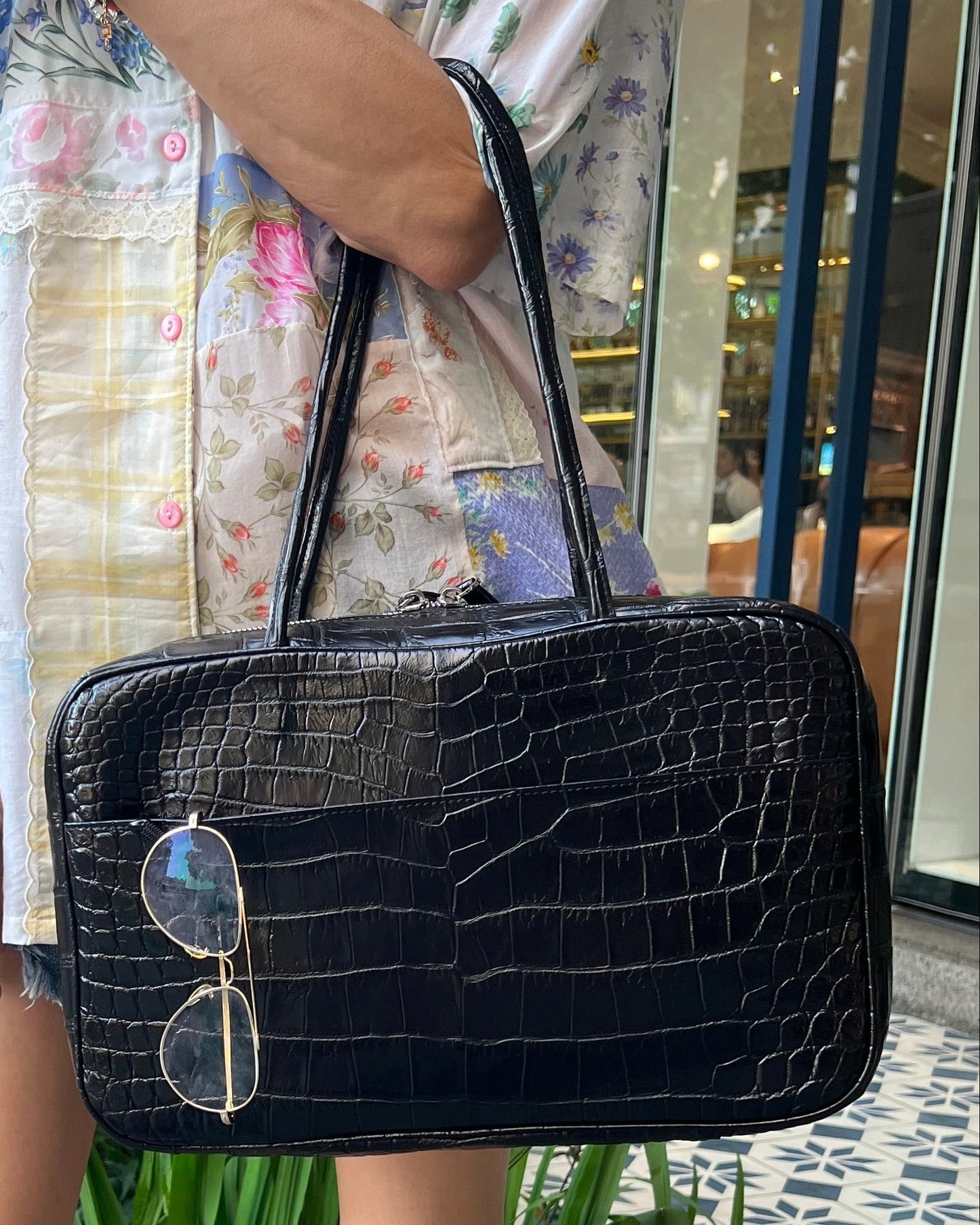 Person holding a black crocodile-patterned handbag with sunglasses on a blurred indoor background