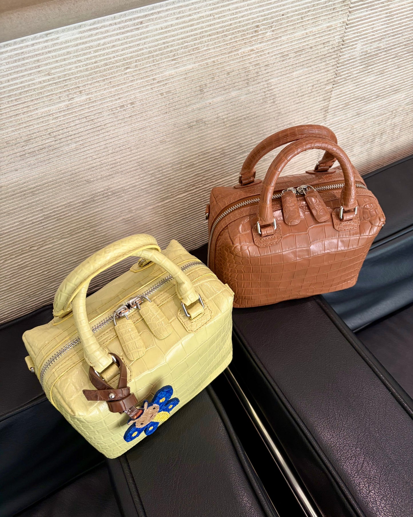 Two structured leather handbags placed on a black leather seat against a textured beige wall. The bag in the foreground is pale yellow crocodile-embossed leather with silver hardware and a blue-and-brown hanging charm.