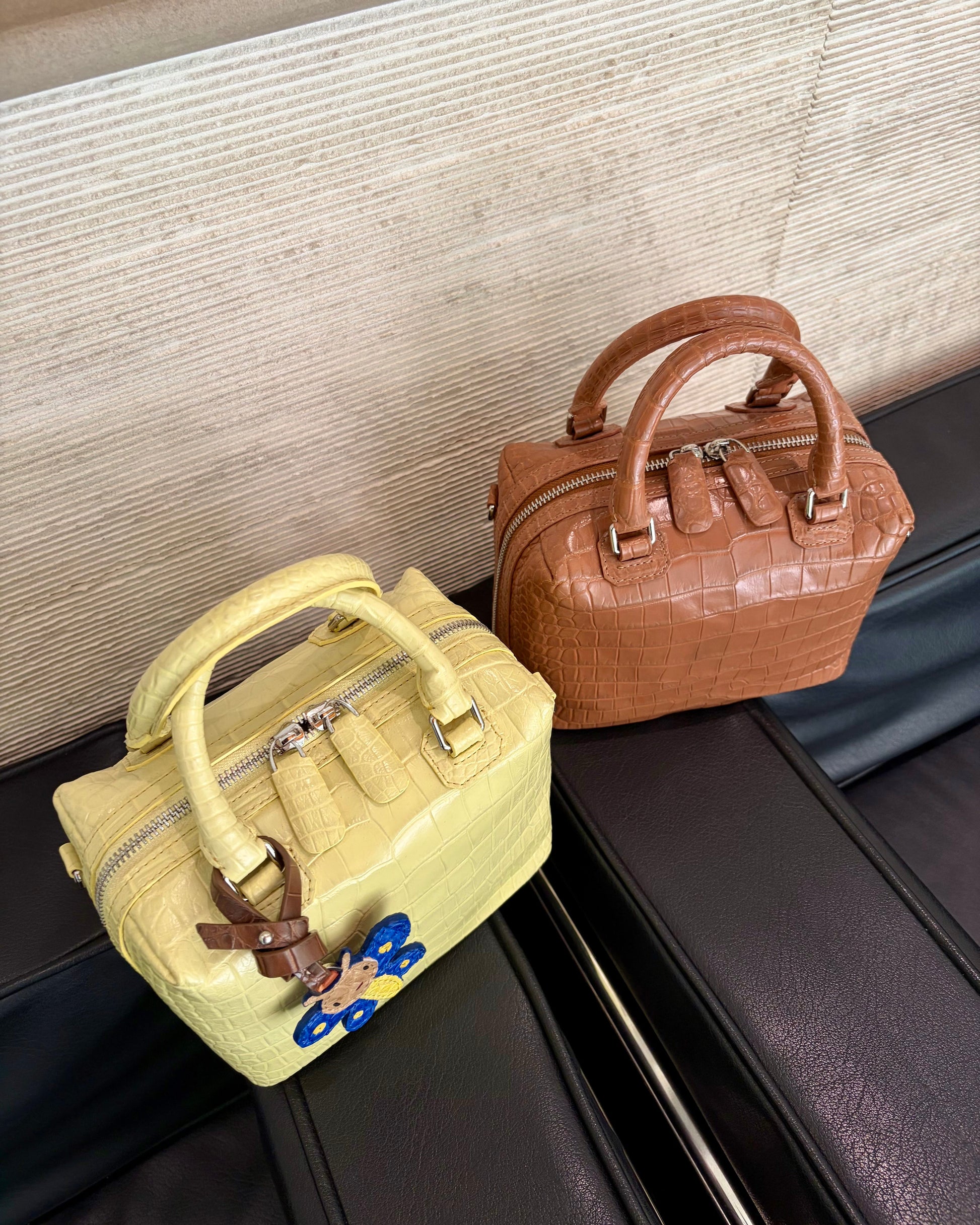 Two structured leather handbags placed on a black leather seat against a textured beige wall. The bag in the foreground is pale yellow crocodile-embossed leather with silver hardware and a blue-and-brown hanging charm.