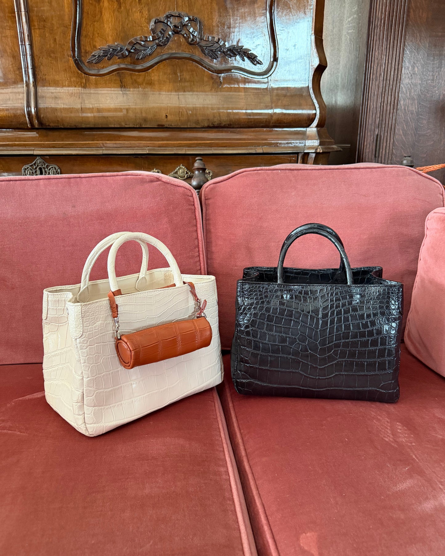 Two designer handbags, one cream crocodile-embossed tote with a brown leather cylindrical pouch and one black crocodile-embossed tote, displayed on a dusty pink velvet sofa in front of a wooden cabinet.